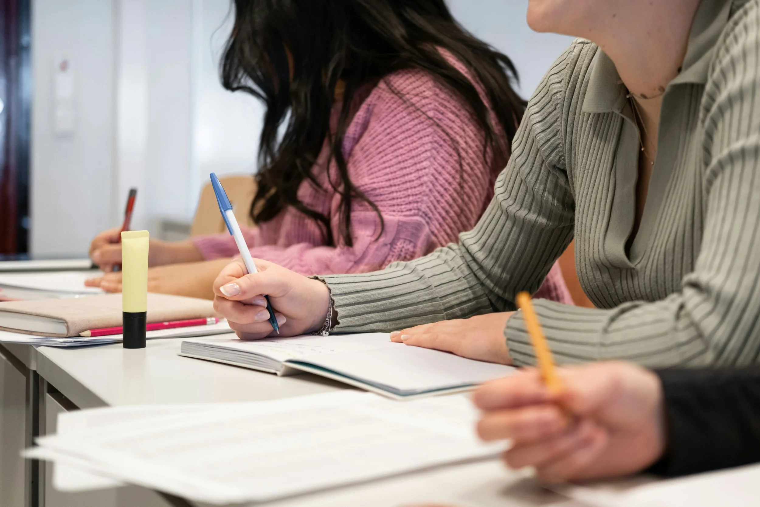 People sitting at a desk, taking notes in notebooks with pens in a classroom or meeting setting.