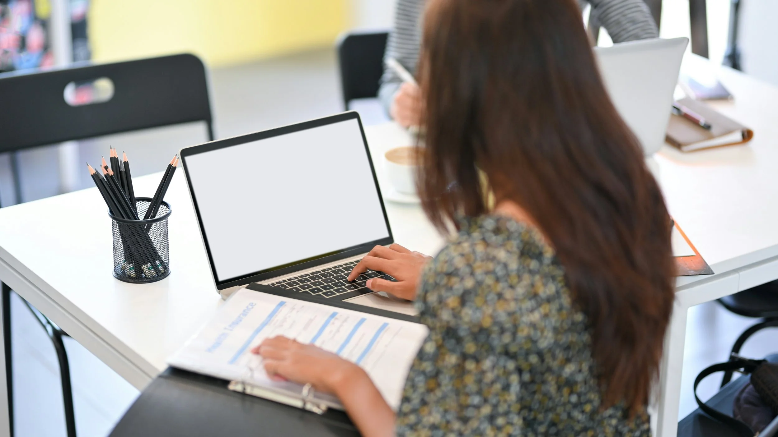 Woman with long brown hair working on a laptop at a white table in a bright, modern workspace.