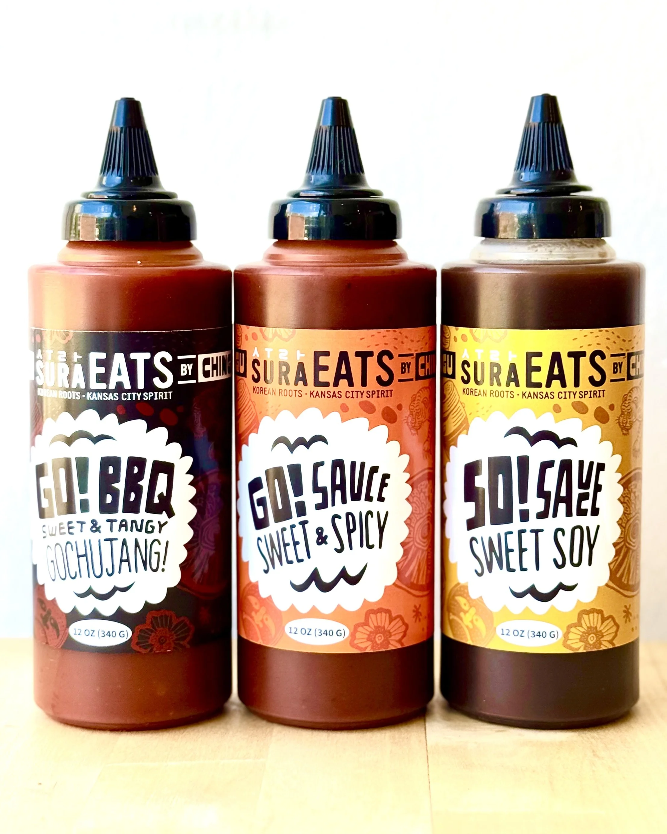 Three bottles of Sura Eats sauces with different flavors, including Sweet & Tangy, Sweet & Spicy, and Sweet Soy. Each bottle has a colorful label and a black squeeze cap, and they are placed side by side on a light surface against a plain background.