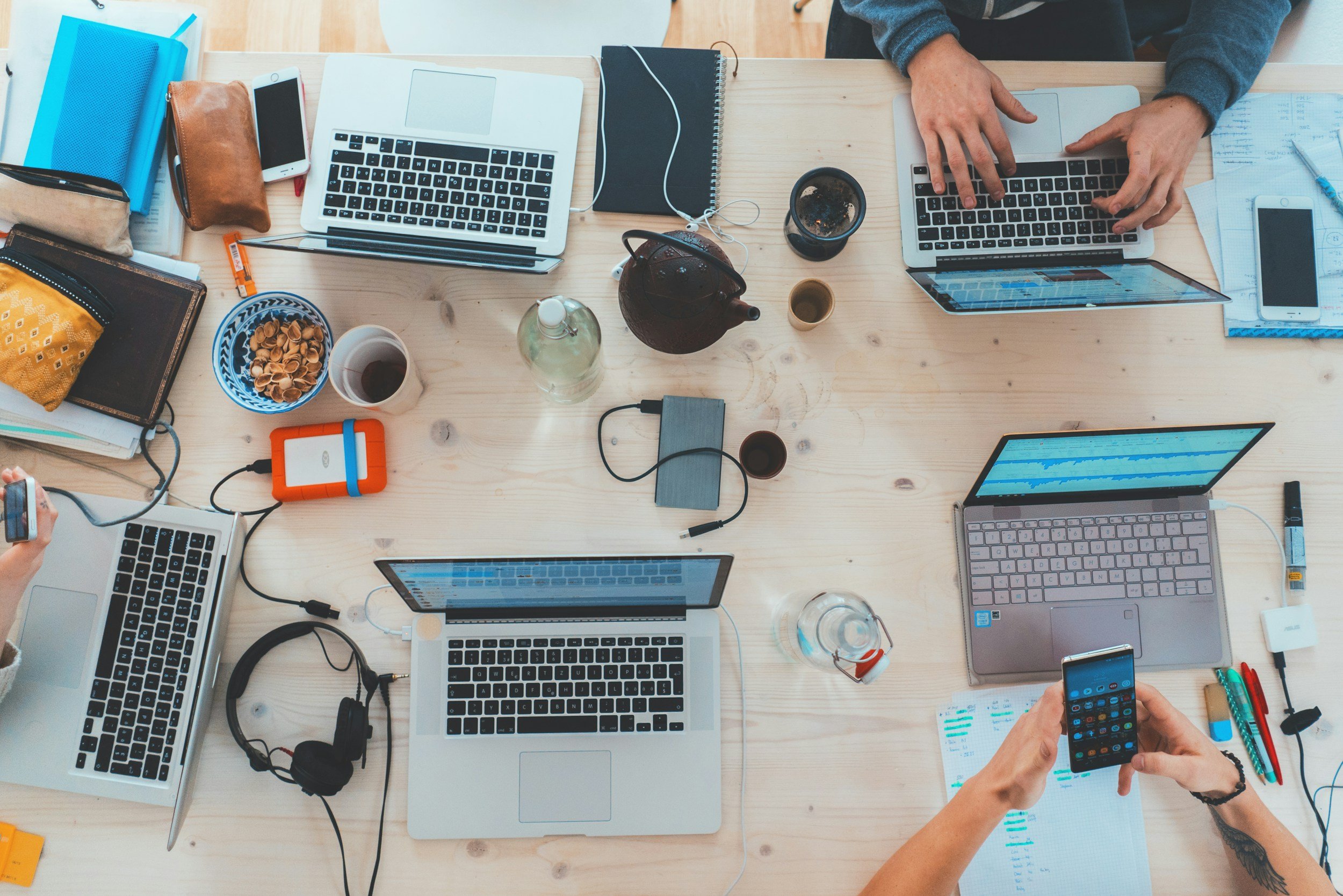 Top-down view of a cluttered wooden desk with multiple laptops, smartphones, notebooks, cups of coffee, and various personal items, with two people working on the laptops.