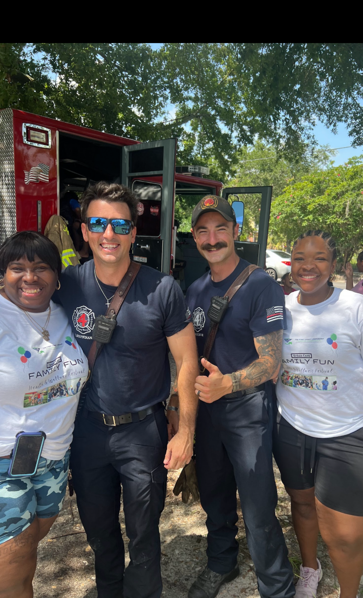Four smiling people standing outdoors in front of a fire truck, participating in a community event. Two women on the ends are wearing white T-shirts with the text 'FAMILY FUN' and colorful graphics, while the two men in the middle are wearing navy blue uniforms with fire department logos. The group is diverse, with a background of green trees and parked cars under a sunny sky.
