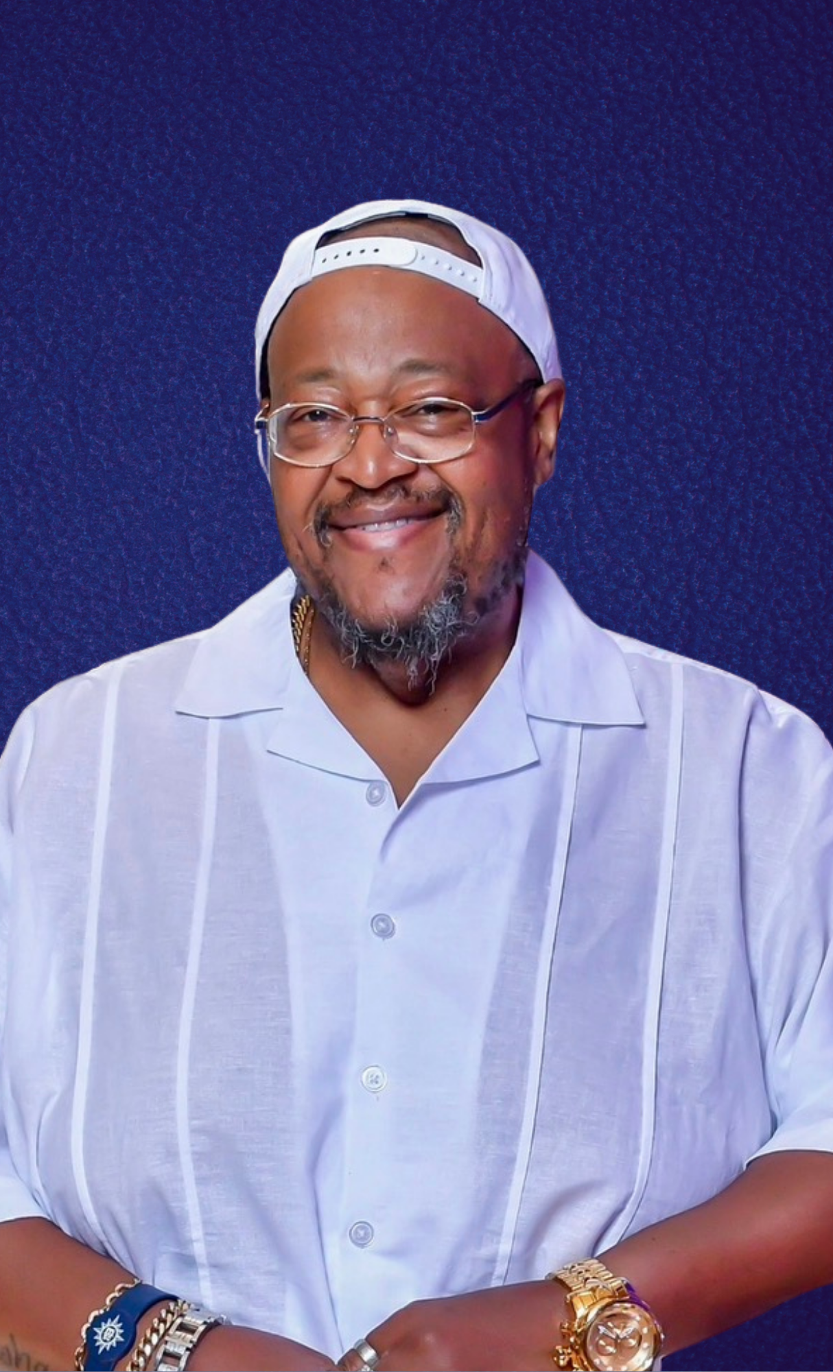 Man smiling, wearing glasses, a white baseball cap backward, a white button-up shirt, and gold jewelry, including watches and bracelets, against a blue background.