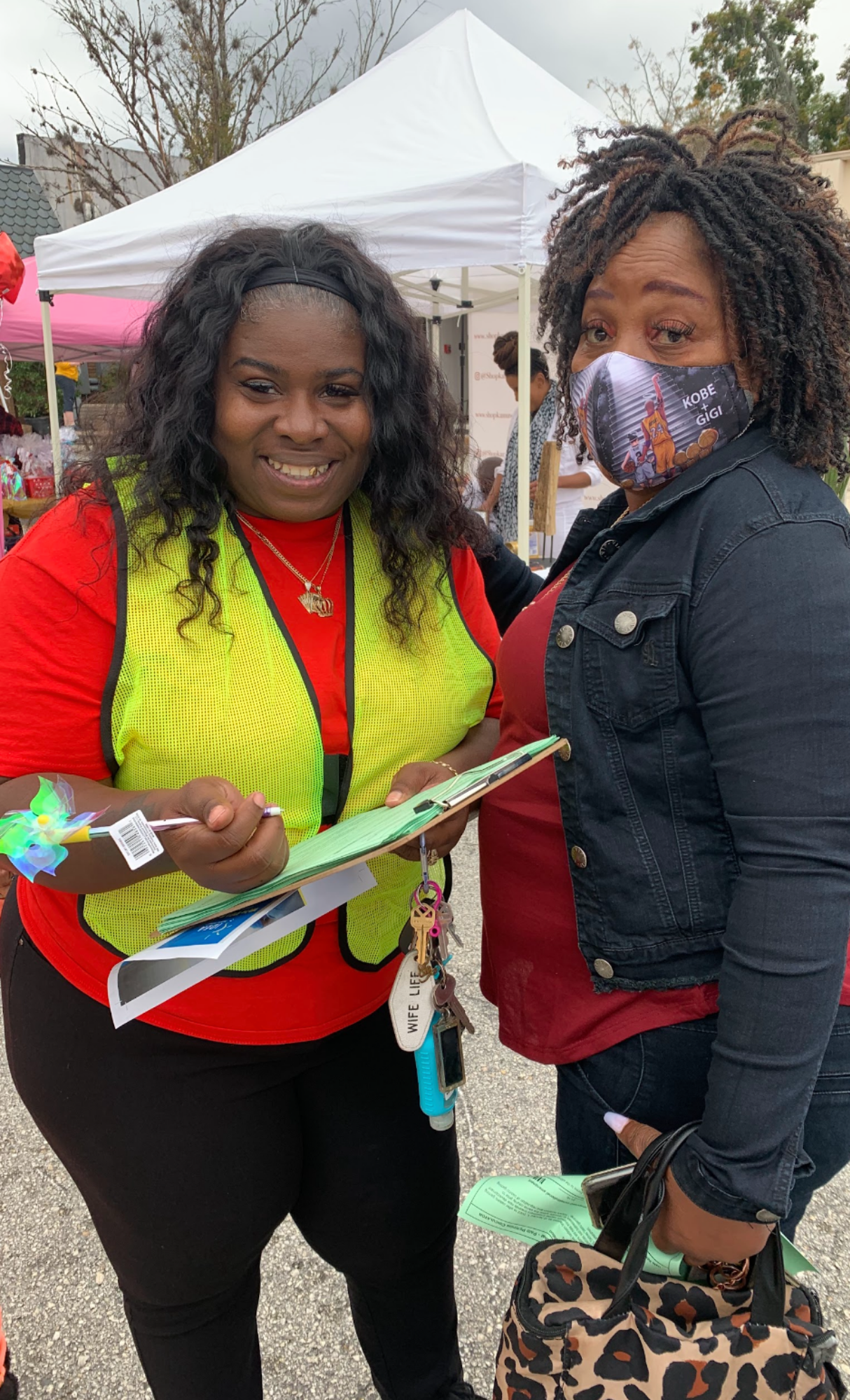 Two women stand facing each other at an outdoor event. One woman wears a yellow vest over a red shirt and holds a clipboard, while the other woman wears a face mask and a black jacket over a red top, holding a leopard print bag. Tents and other people are visible in the background.