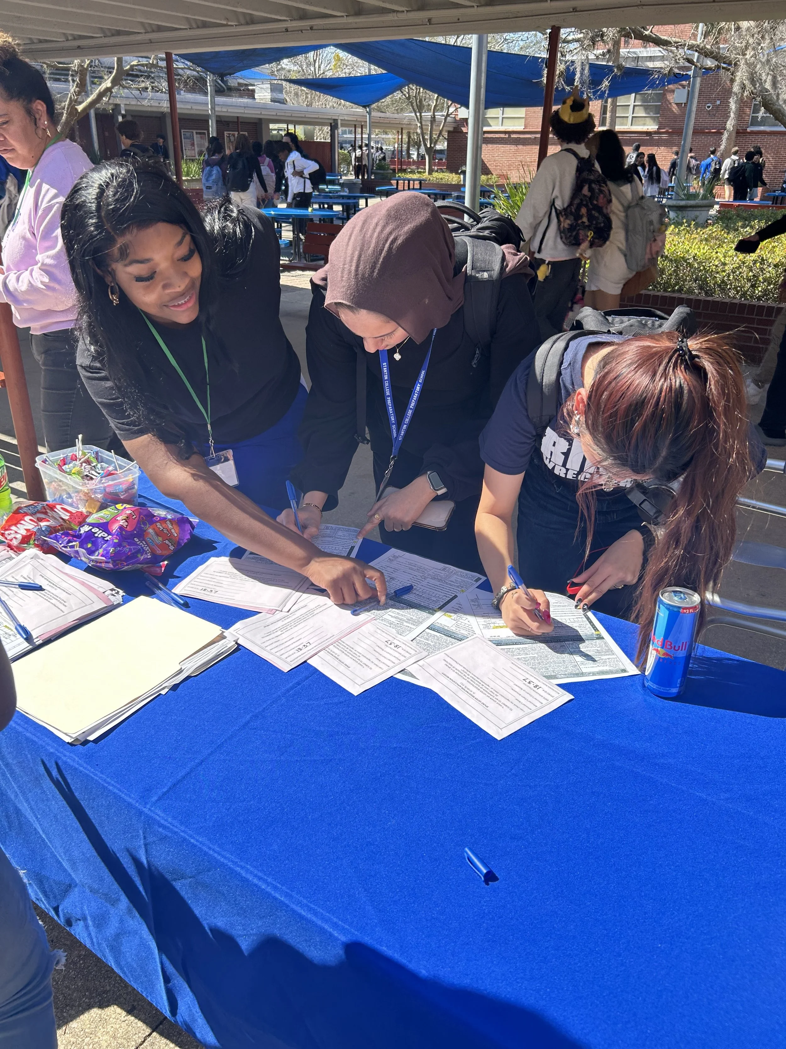 People filling out paperwork at a table covered with a blue cloth outdoors, with snacks and a Red Bull energy drink on the table, in a school courtyard on a sunny day.