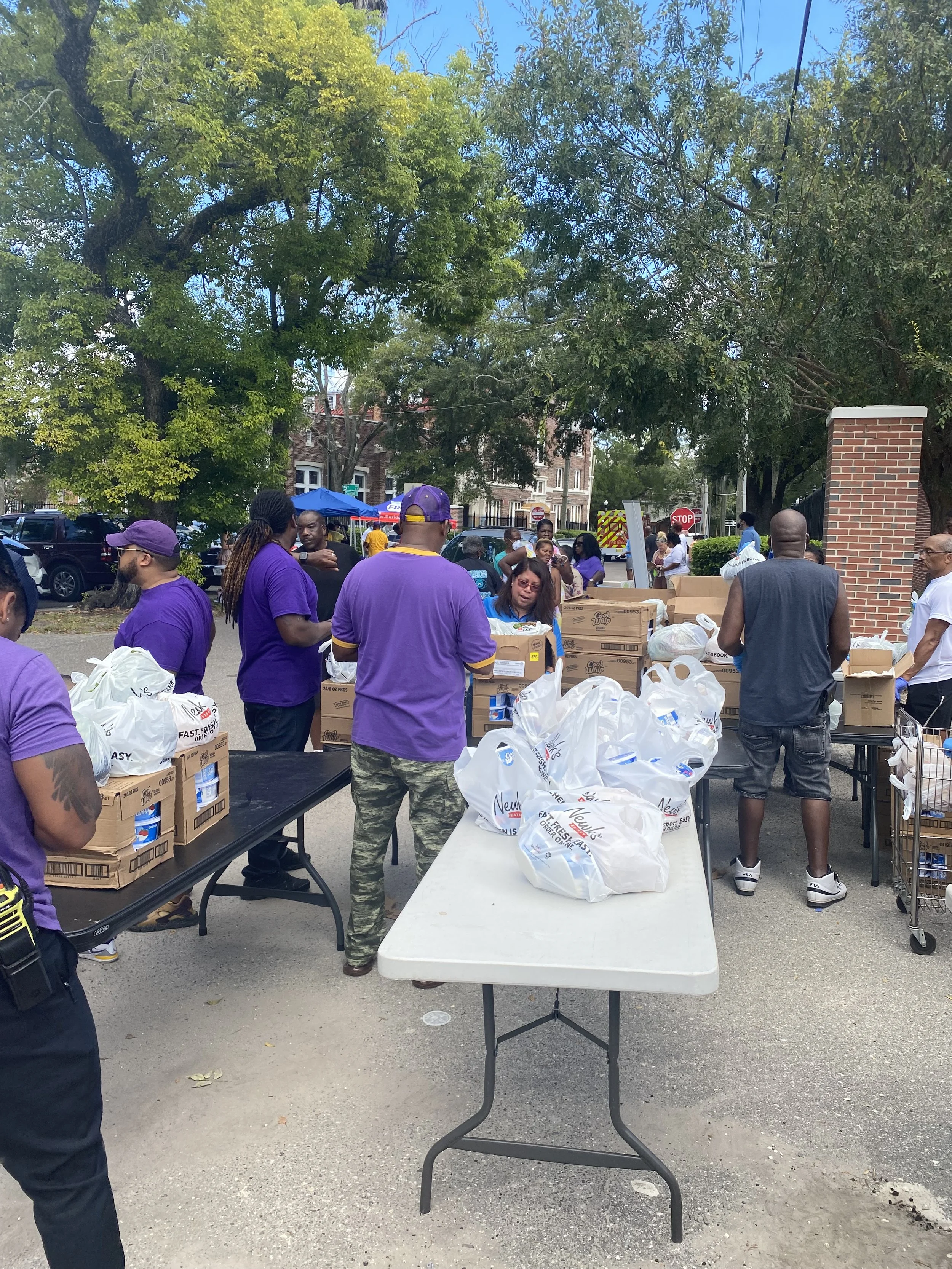 People distributing food and supplies outdoors on a sunny day, with trees and buildings in the background.