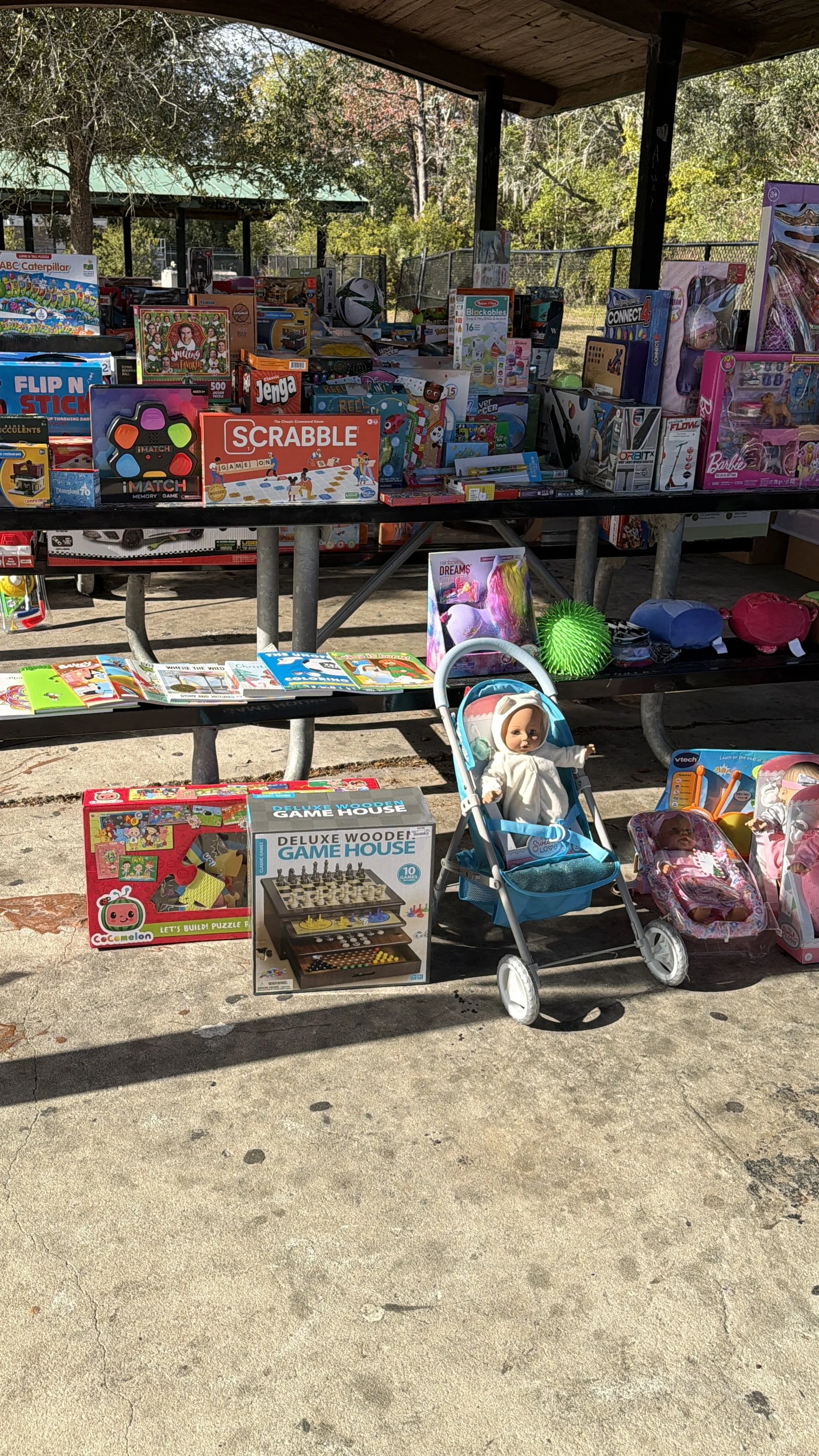 Outdoor sale of toys including board games, dolls, and stuffed animals on a metal picnic table with a concrete ground. Items include a doll in a stroller, toy chess set, baby doll, and model trains, among others. Trees and a picnic shelter are in the background.