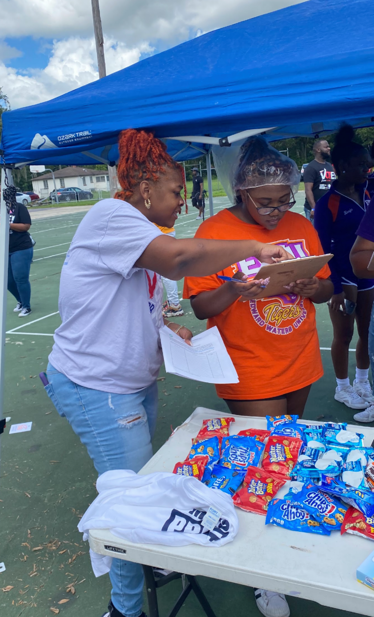 Two women standing at a table of assorted snack bags outdoors under a blue canopy, with a basketball court and other people in the background. One woman with curly red hair, wearing a white T-shirt, is pointing to something on the clipboard held by the other woman, who is wearing glasses, an orange T-shirt, and a plastic hair cap.