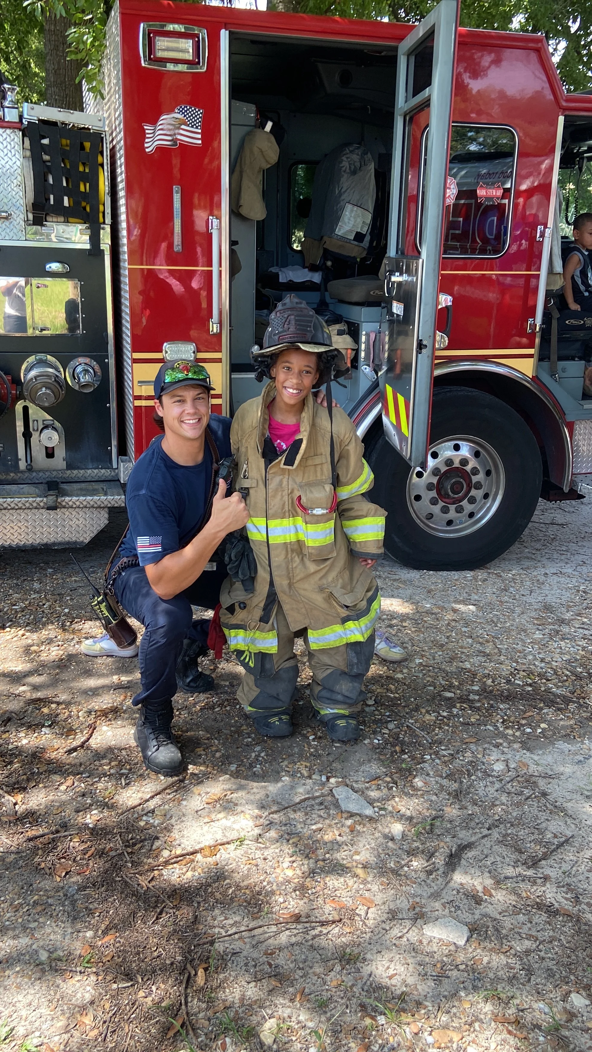 A firefighter and a young girl smiling in front of a red fire truck, with the girl dressed in firefighter gear and the firefighter giving a thumbs-up.