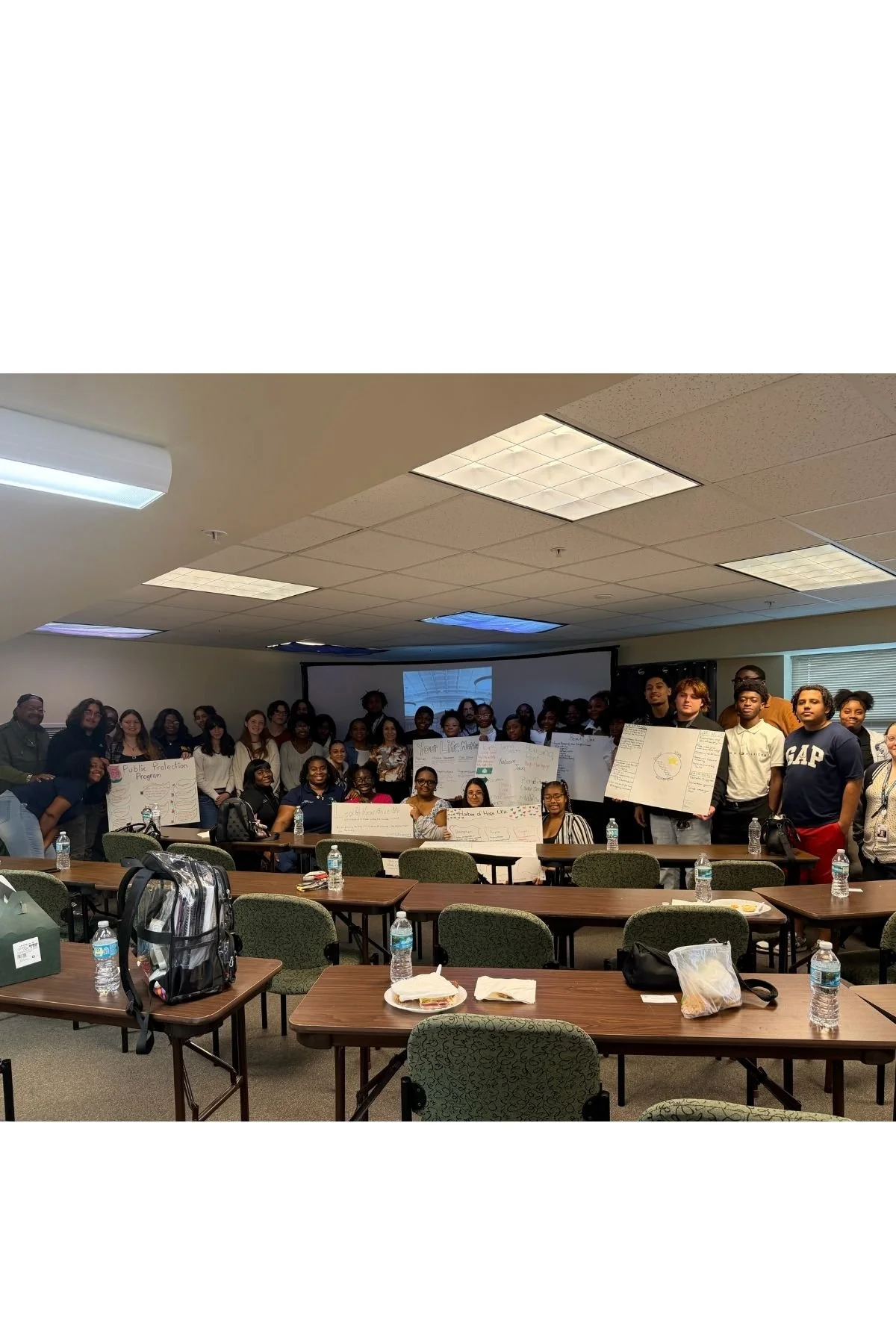 Group of diverse young people gathered in a classroom or conference room, holding posters and smiling for a group photo.