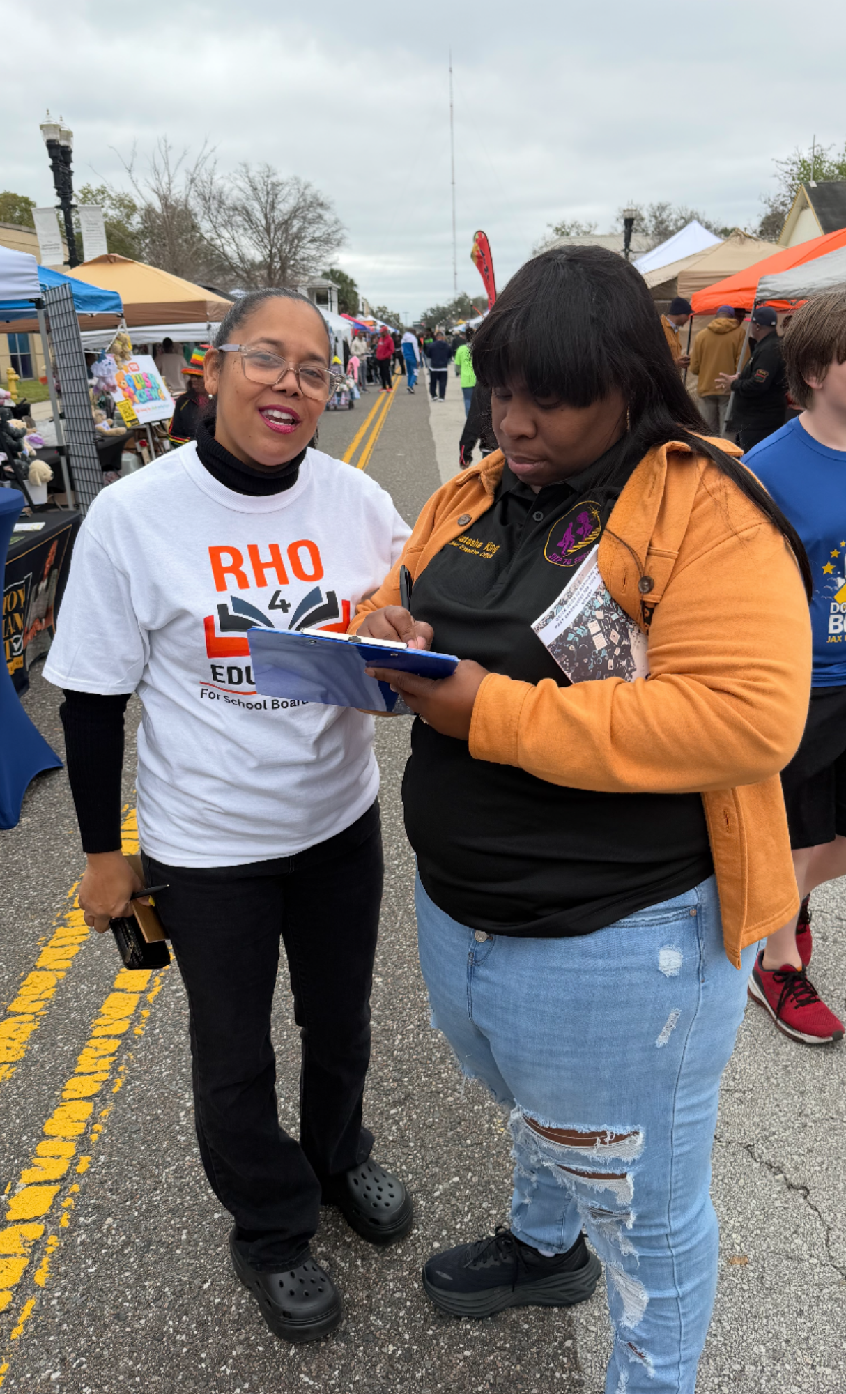 Two women standing in the middle of a crowded outdoor street fair, with tents and vendors on either side. One woman is wearing a white shirt that says "RHO 4 EDUCATION For School B" and is holding a phone, while the other woman is wearing a black shirt with a purple emblem and a brown jacket, and is writing on a clipboard. Overcast sky.