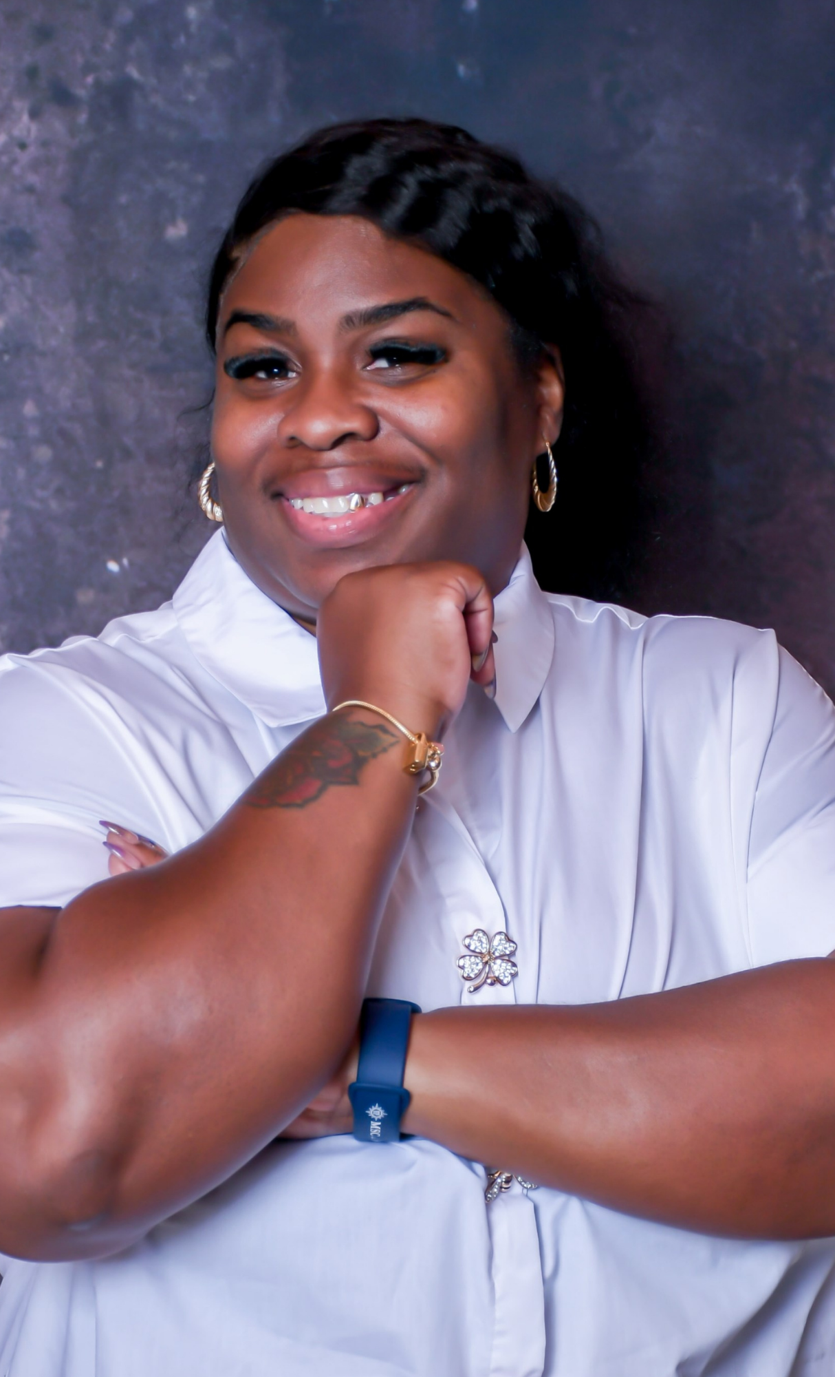 portrait of a smiling woman with dark hair, wearing a white shirt, angled slightly to the side, with her fist resting near her chin, against a dark, textured background.