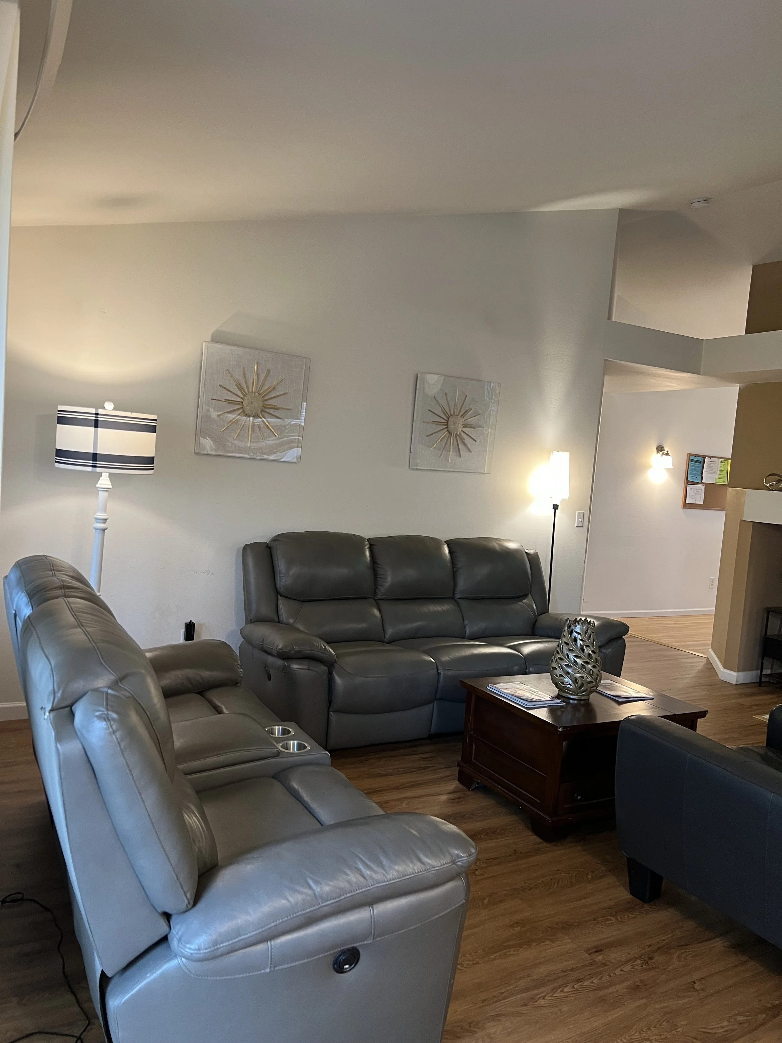 NEIGHBORS AFH INC Living room with gray leather recliner, gray leather sofa, wooden coffee table with decorative vase, wall art, floor lamp, and ceiling lights.