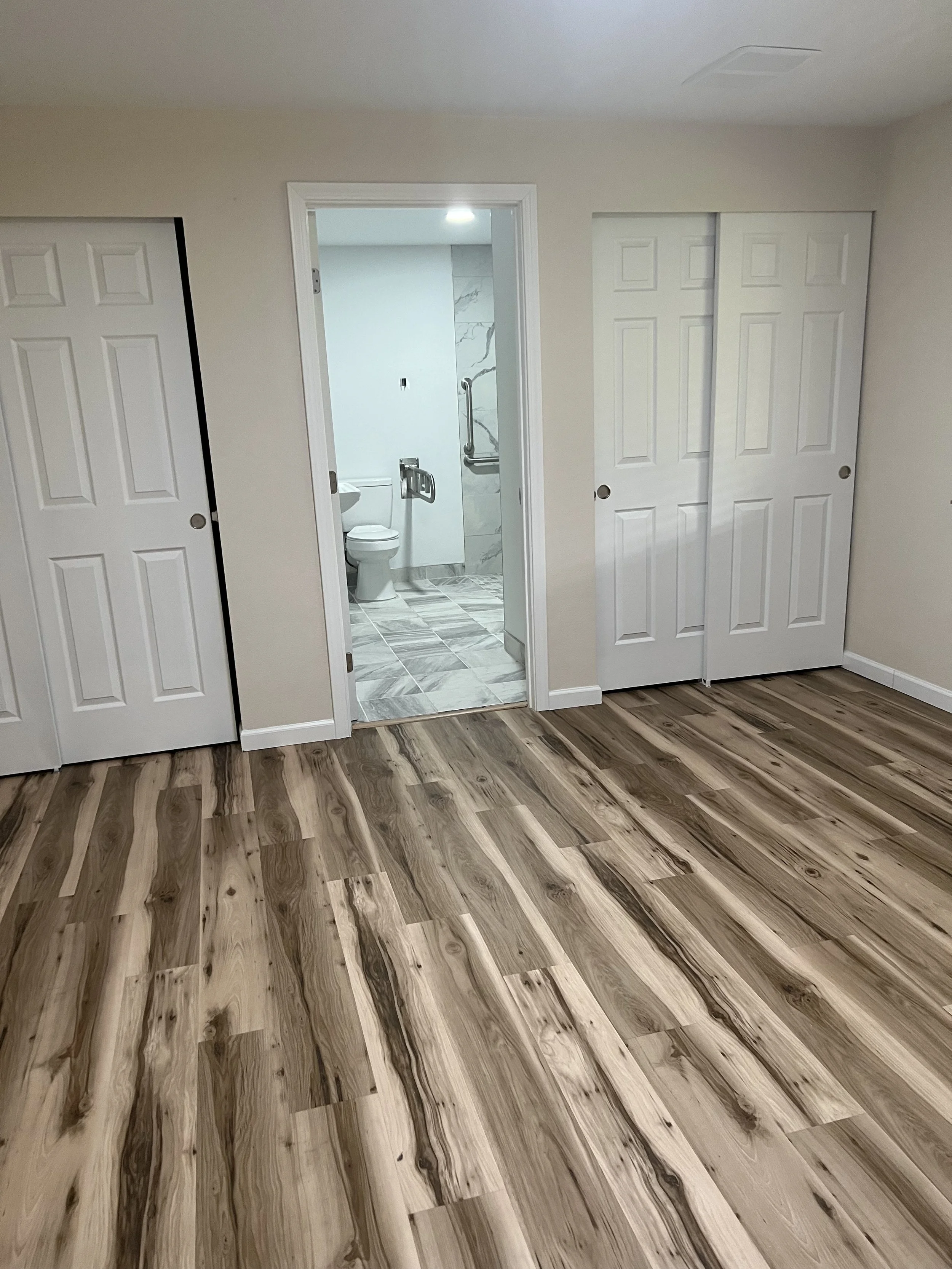 NEIGHBORS AFH INC Interior view of a room with wood flooring, a closed closet door, and an open door leading to a bathroom with marble tiles and a toilet.