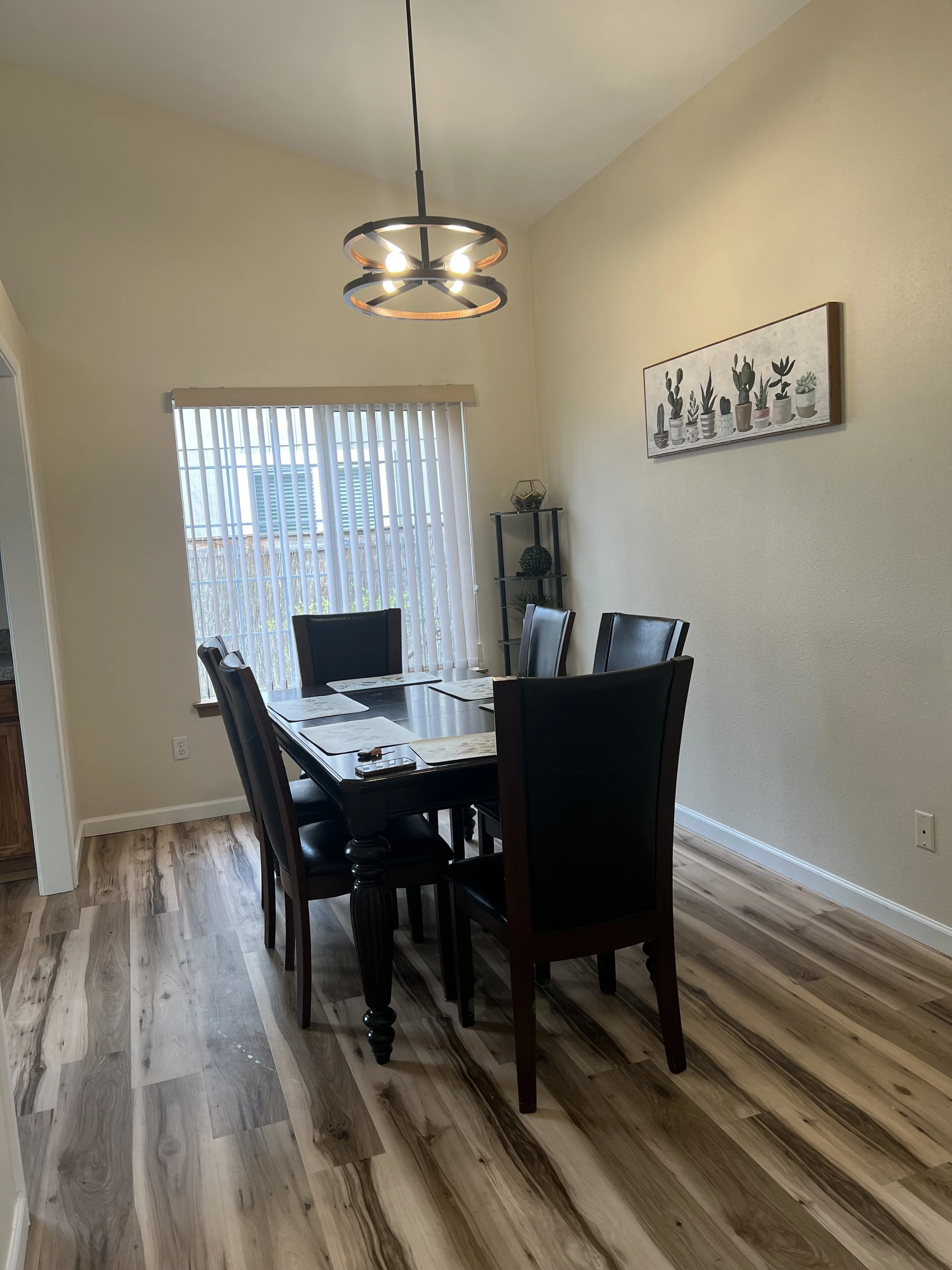 NEIGHBORS AFH INC dining room with a dark wooden table and six matching chairs, a window with vertical blinds, a light-colored wall with a framed picture of various potted plants, and a ceiling light fixture with circular rings and exposed bulbs.