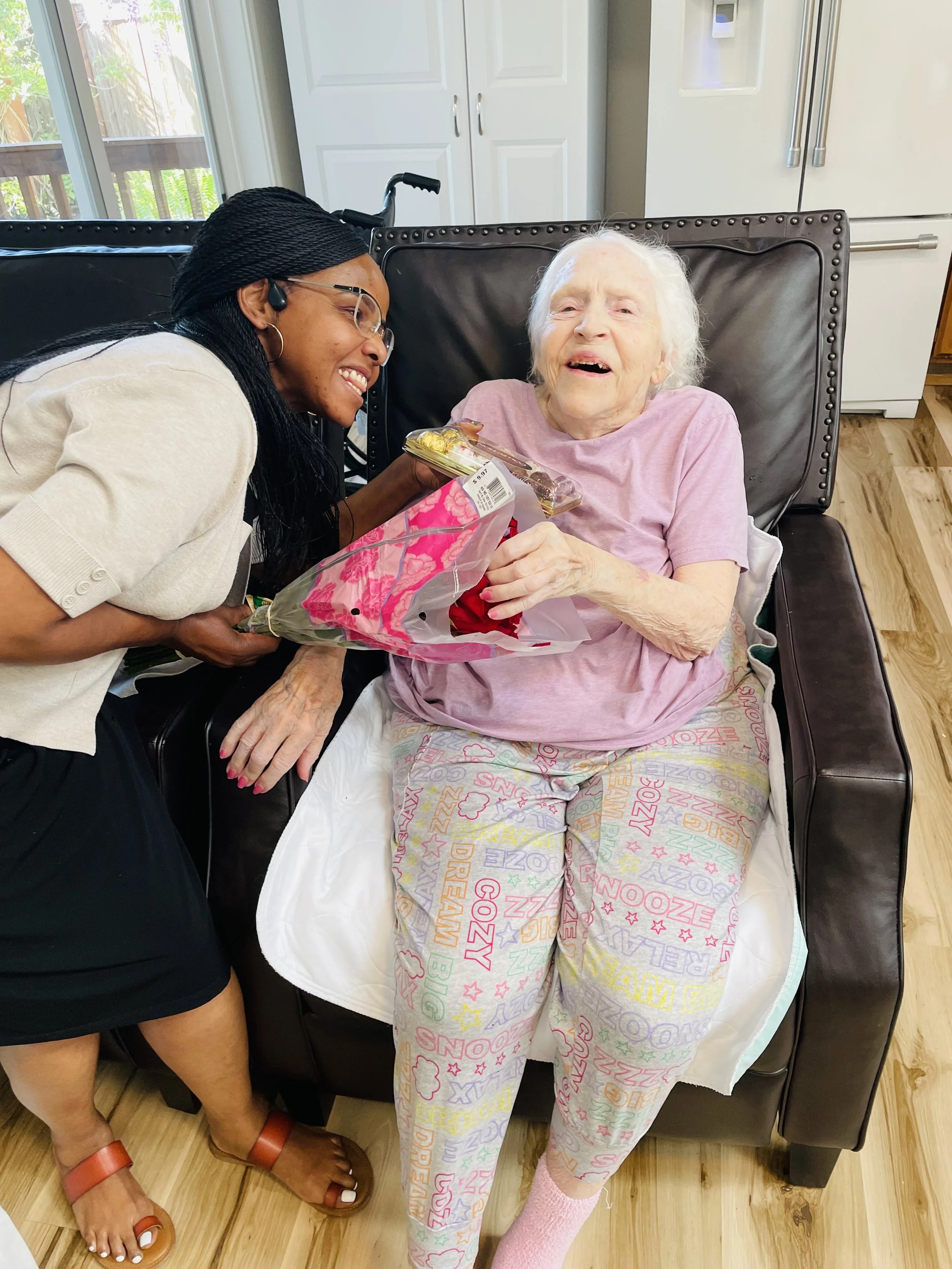 An elderly woman with white hair sitting in a wheelchair, smiling and holding a pastry. A young woman, wearing glasses and earrings, is leaning in happily, holding a bouquet of flowers towards the elderly woman. The scene takes place in a bright kitchen with a door and fridge in the background.