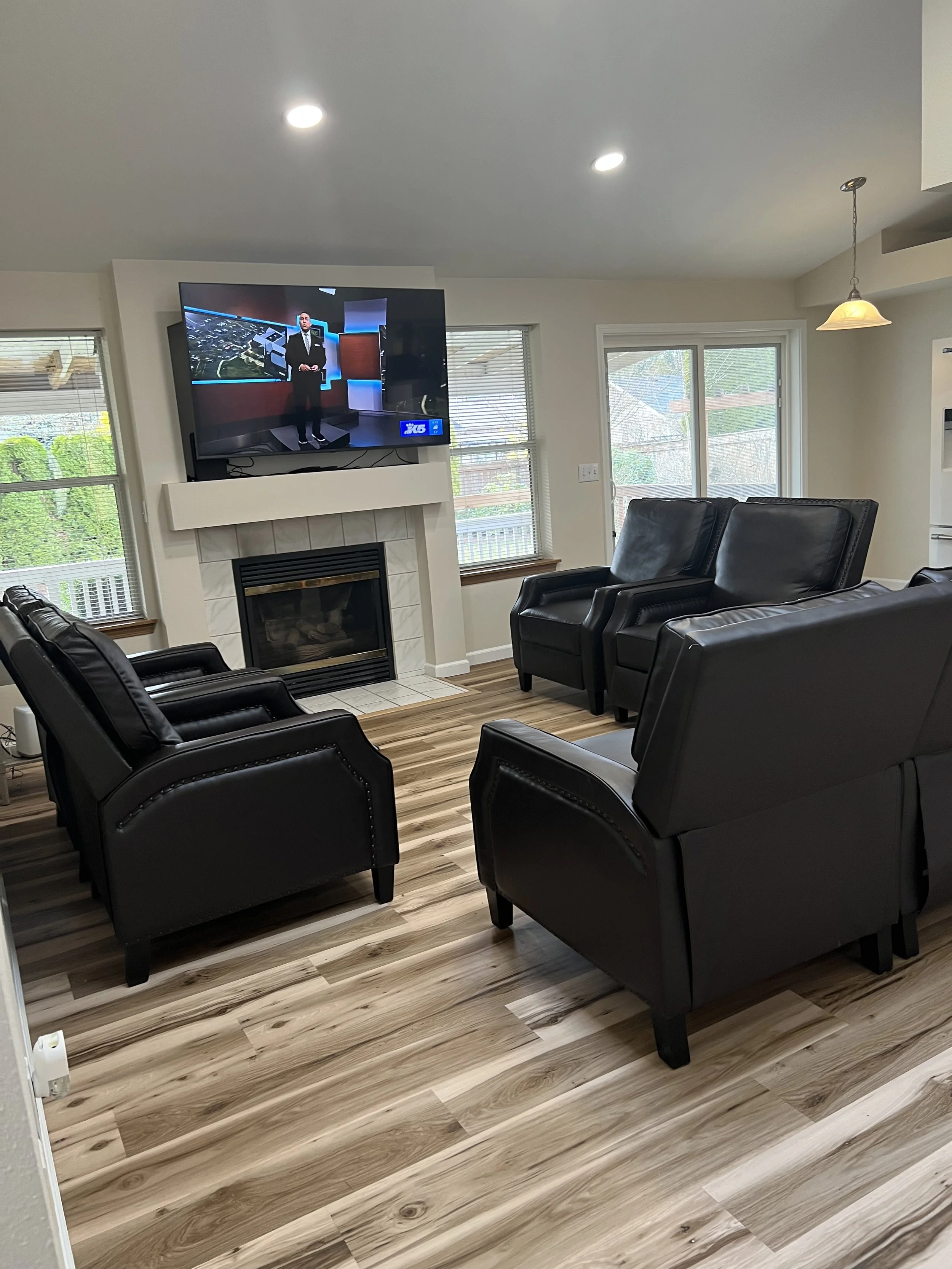 NEIGHBORS AFH INC Living room with black leather armchairs facing a television mounted above a white fireplace.