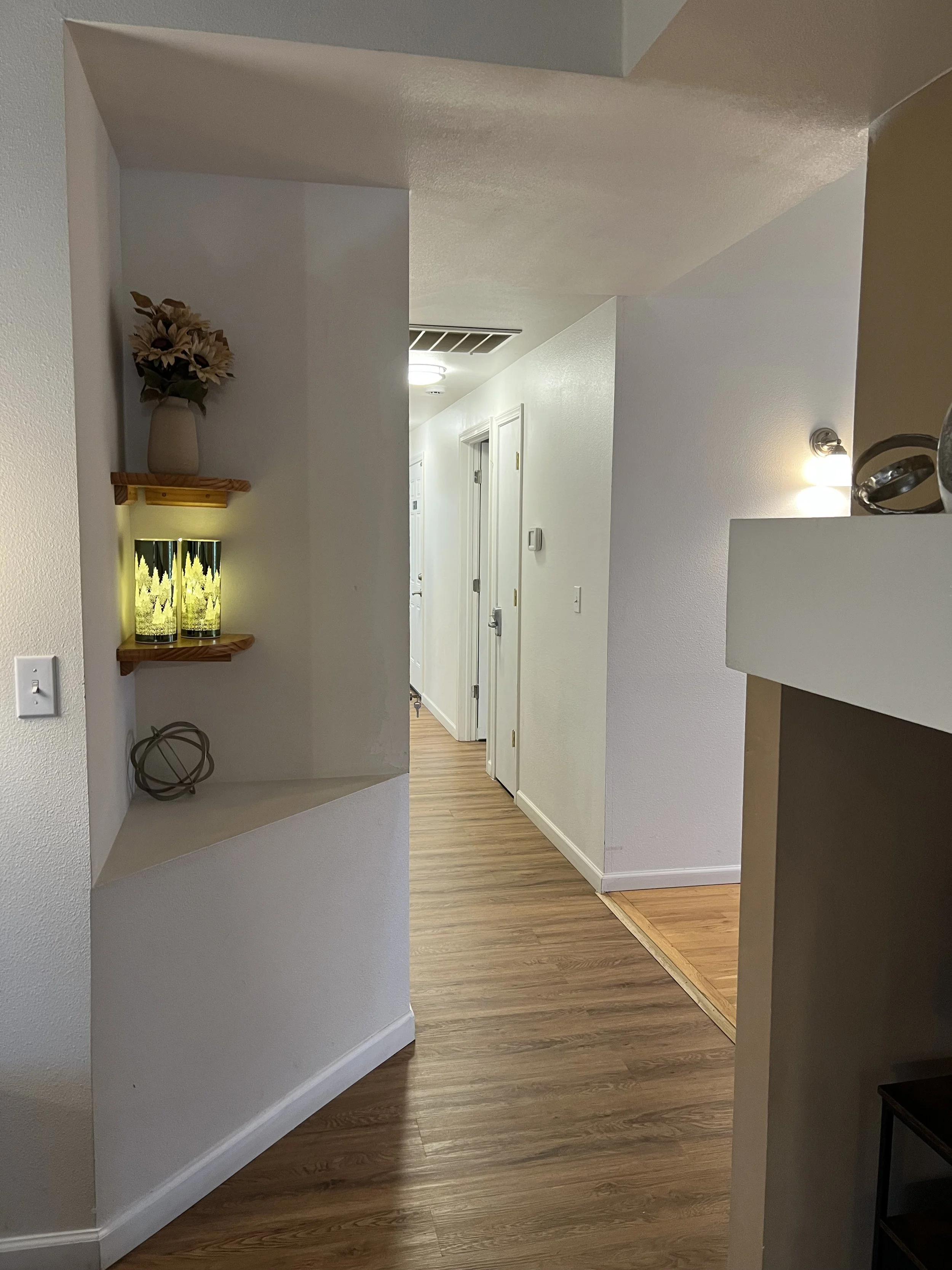 NEIGHBORS AFH INC Interior hallway with wooden flooring, white walls, a small wall shelf with a vase of flowers, two yellow decorative lights, and a partially visible kitchen or living room area.