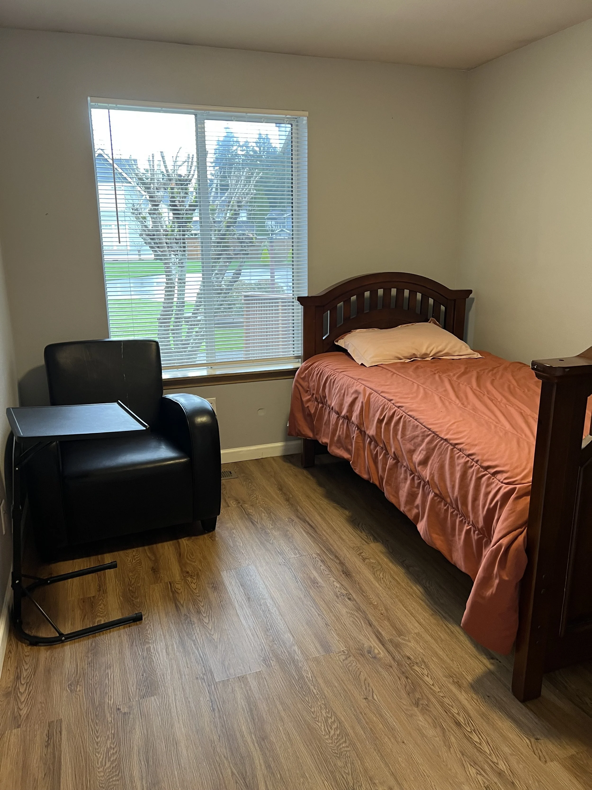 NEIGHBORS AFH INC A bedroom with wooden flooring, a bed with an orange comforter and a pillow, a black desk chair, and a window with blinds showing trees outside.