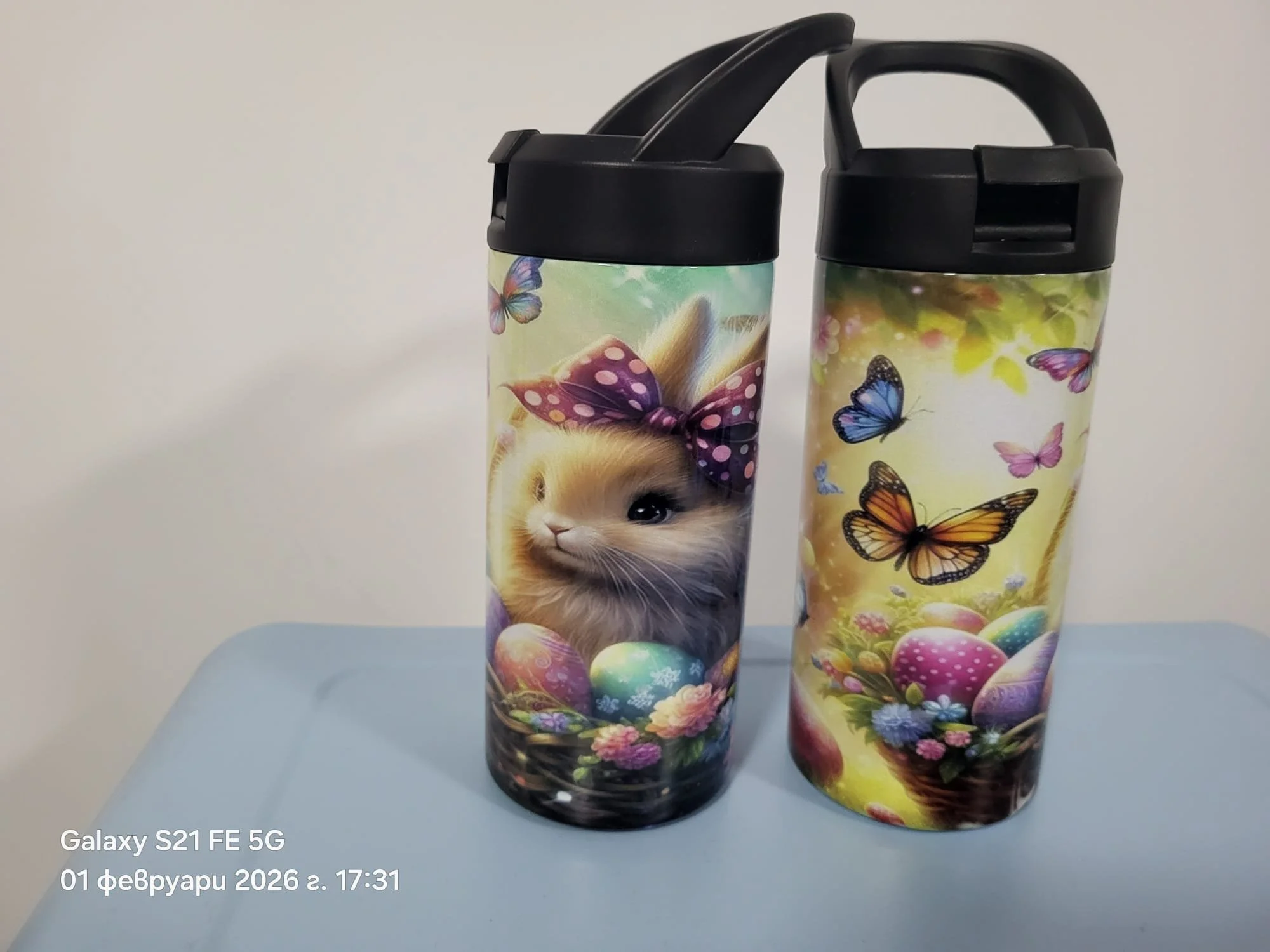 Two colorful travel mugs with black lids and handles, decorated with images of puppies and butterflies, placed on a light surface against a plain background.