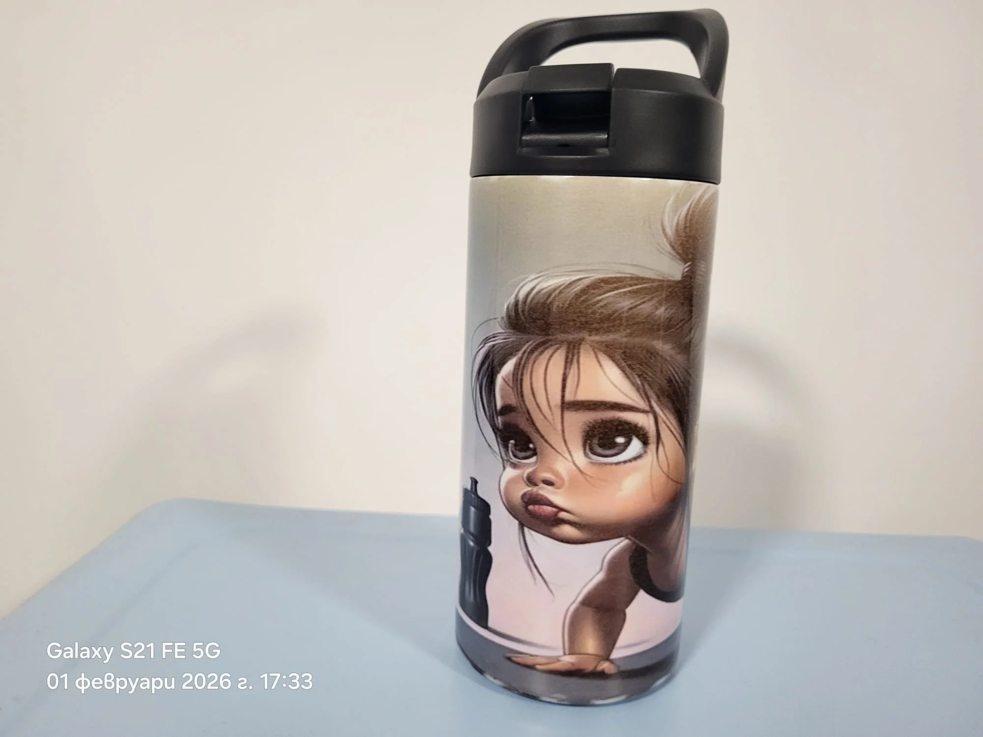 A picture of a stainless steel tumbler featuring a cartoon illustration of a girl with brown hair and large expressive eyes, positioned vertically on the tumbler. The background is plain and light-colored.