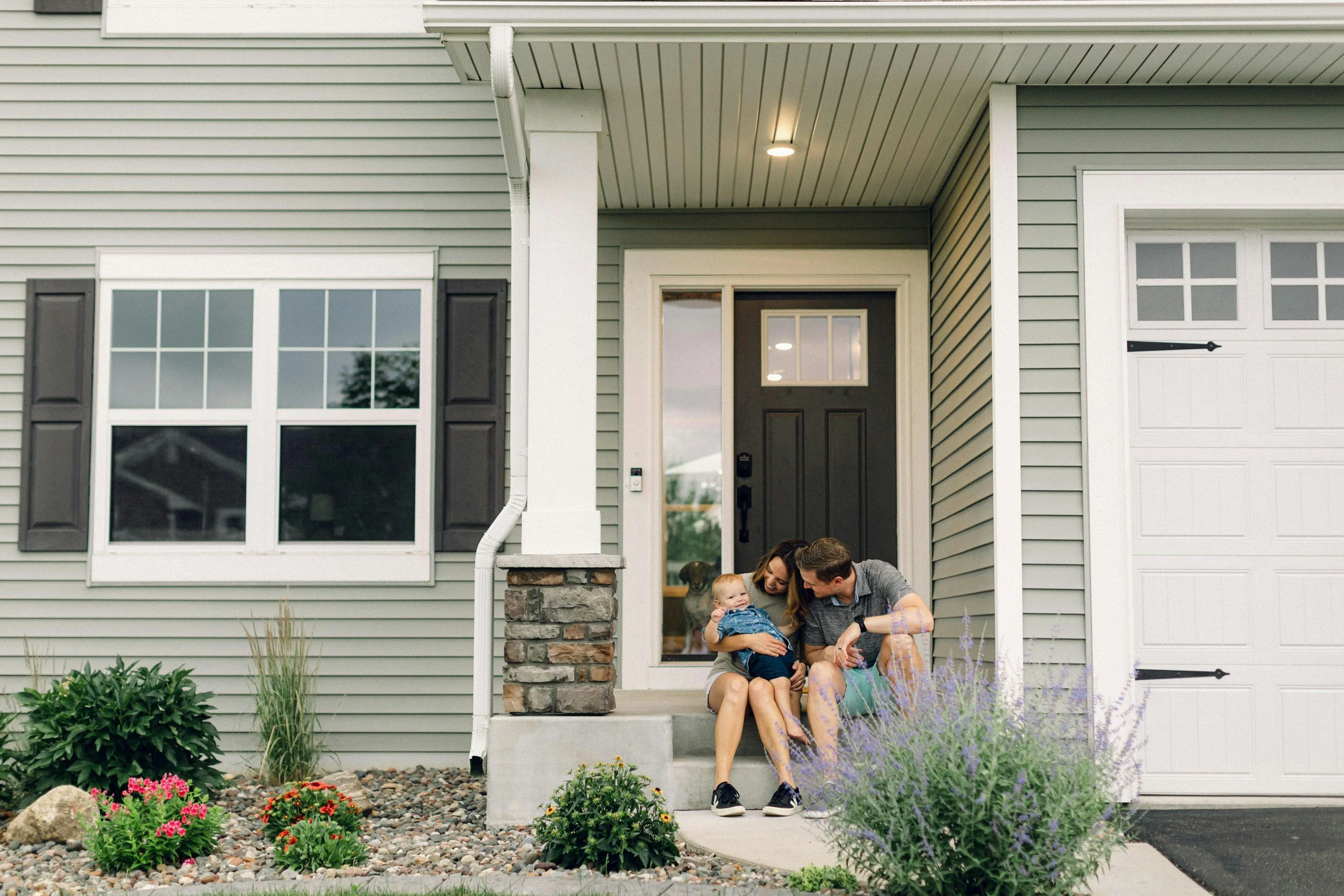 A family sitting on the front porch of a house, with a woman holding a young child and a man sitting beside them. There is a dog behind them in the house doorway, and the porch is decorated with plants and flowers.