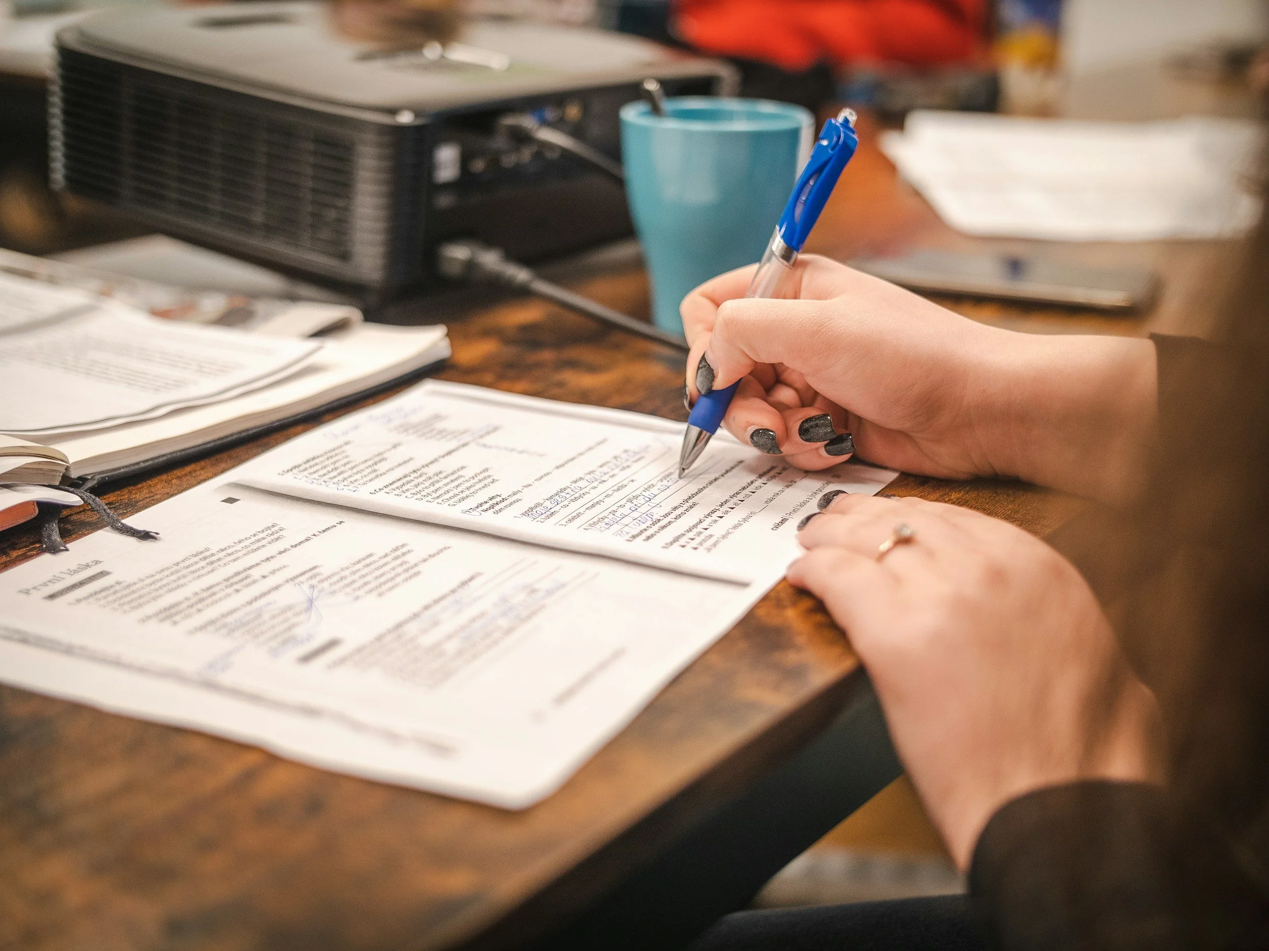 A person is writing on a paper with a blue pen, with office supplies and a blue cup in the background on a wooden desk.