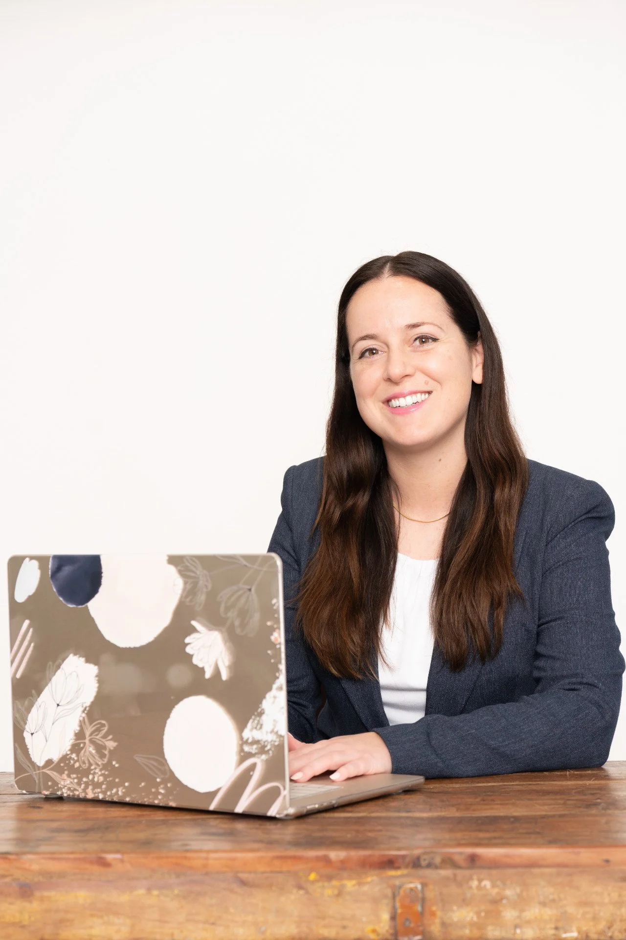 Wendy Brown wearing a blue blazer with a white shirt and dark blazer, sitting at a wooden table with a floral laptop in front of her, smiling against a plain white background.