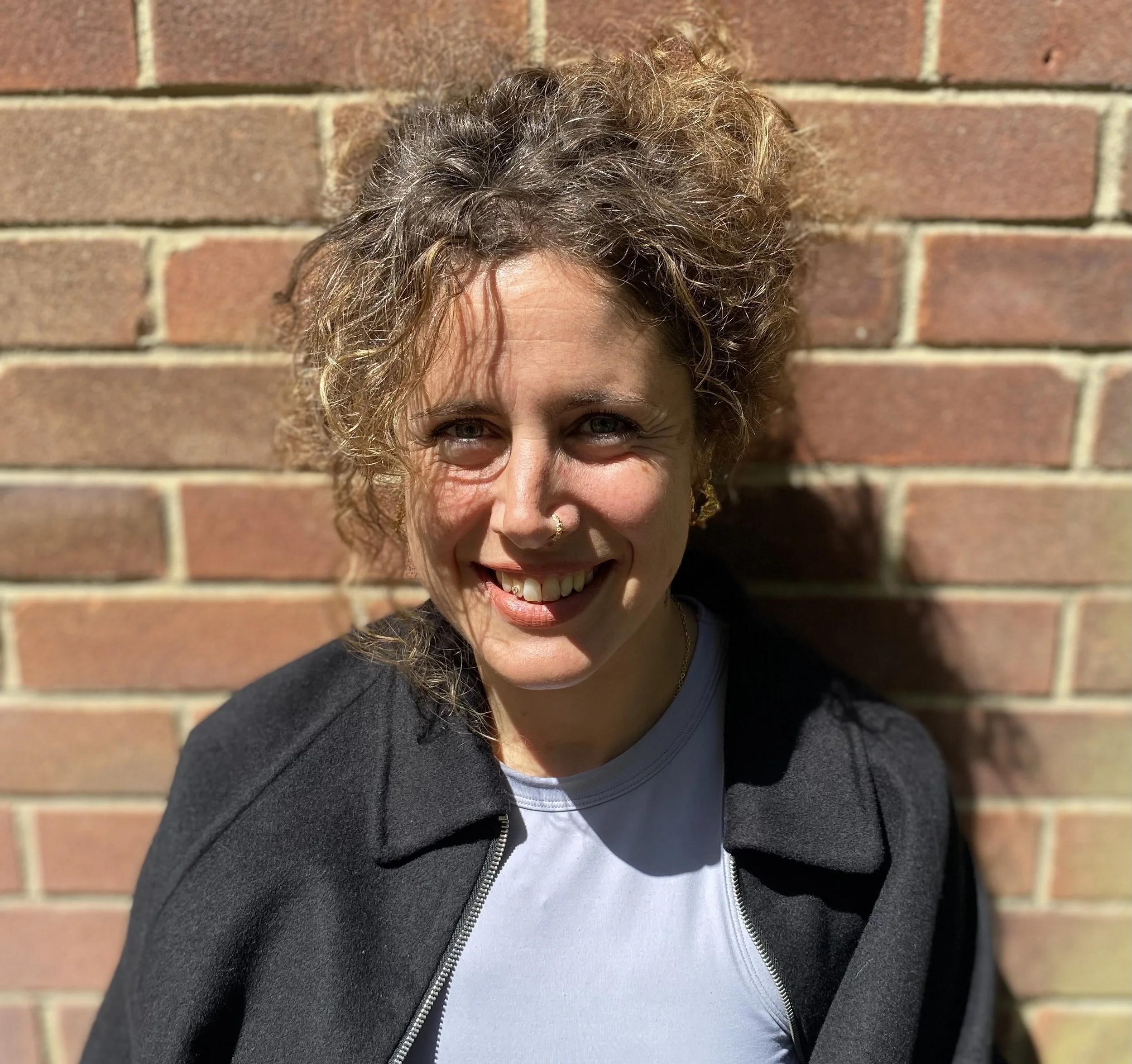 Close-up of a woman with curly brown hair, smiling, standing in front of a brick wall.