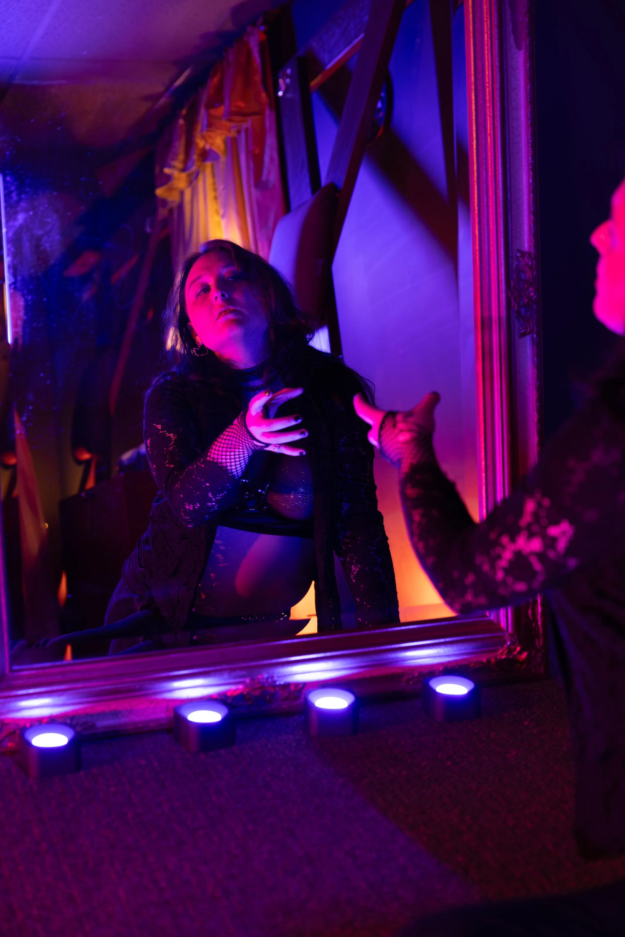 A woman with dark hair and gothic style makeup looks into a reflection in a mirror framed with ornate gold. The room is lit with vibrant purple and pink neon lights, creating a moody atmosphere. The woman is wearing a black lace outfit with fishnet sleeves and has a hand with colorful painted nails touching her chest.