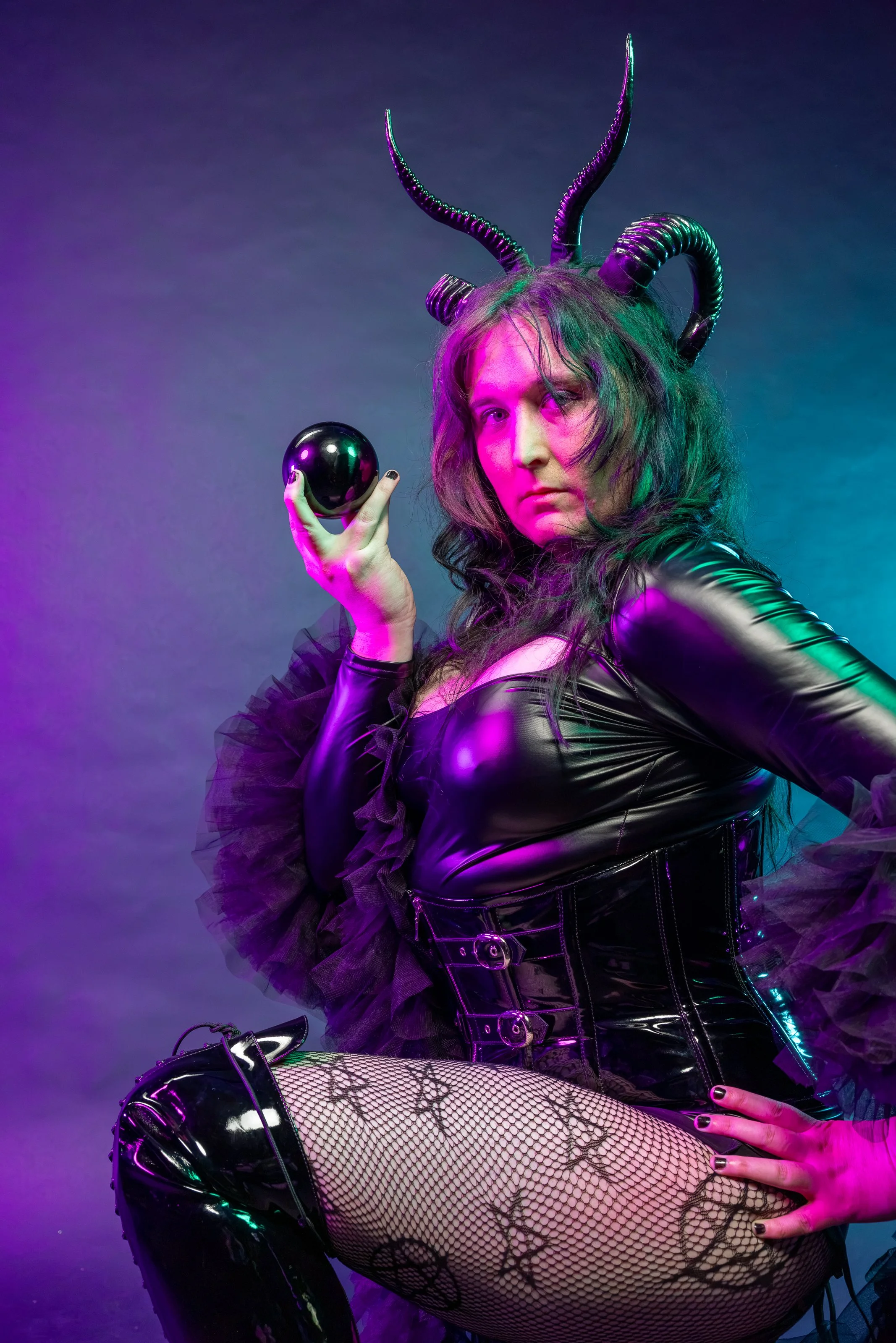 A person dressed in a shiny black leather outfit with tulle ruffles, fishnet stockings with a floral pattern, and black patent boots. They wear a headpiece with black horns and hold a black orb in their left hand, with colorful lighting effects enhancing the scene.