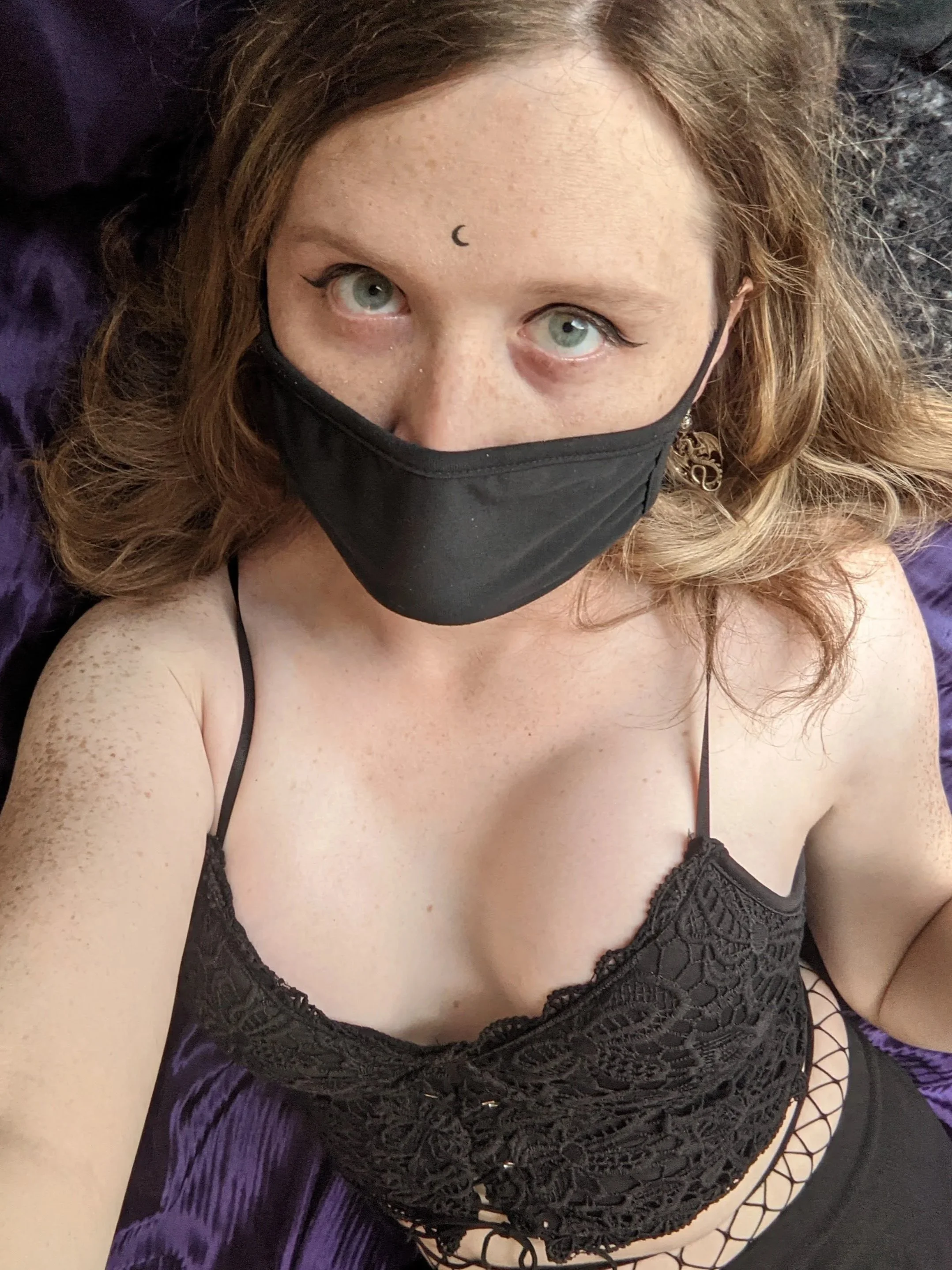 A woman with green eyes and freckles wearing a black face mask and a black lace top, lying on a purple satin surface with black fishnet stockings visible on her arm.