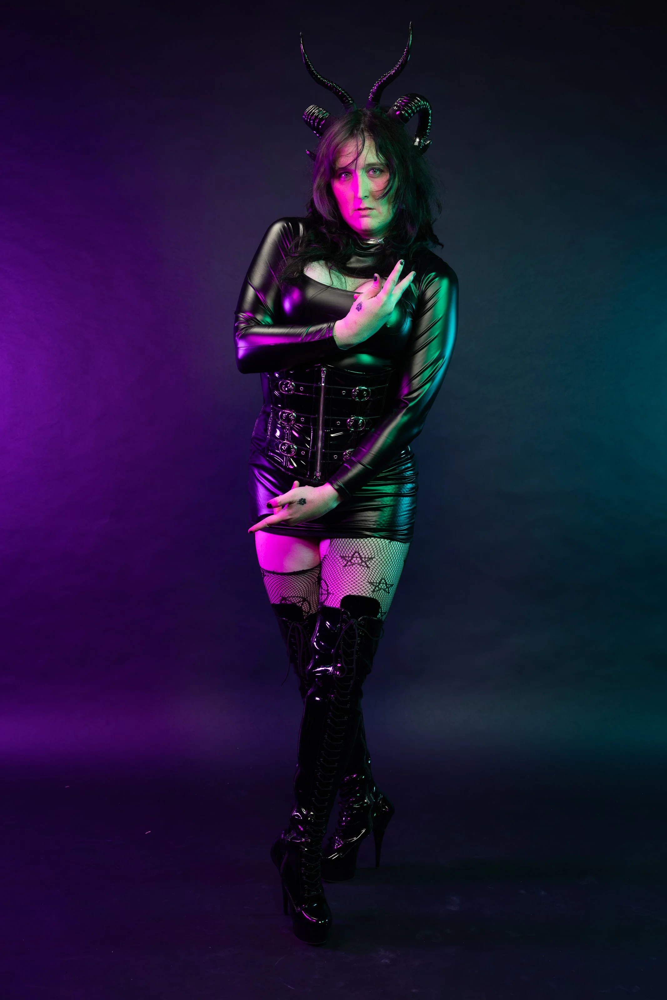 A woman dressed in black latex clothing with horned headpiece, fishnet stockings with star patterns, and high-heeled boots, standing against a dark background with colorful lighting.
