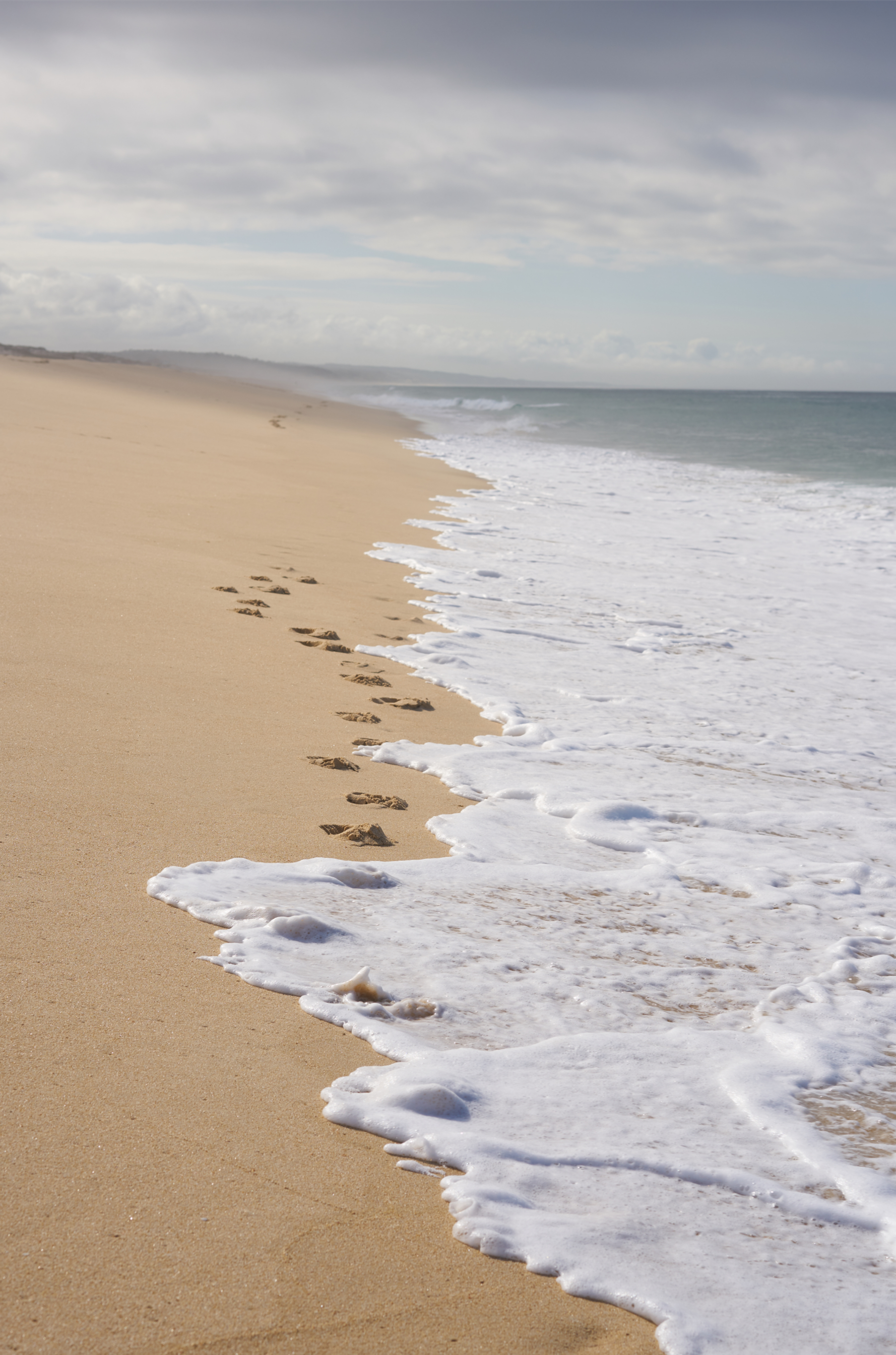 Footprints in the sand along the shoreline where waves are crashing on a cloudy beach.