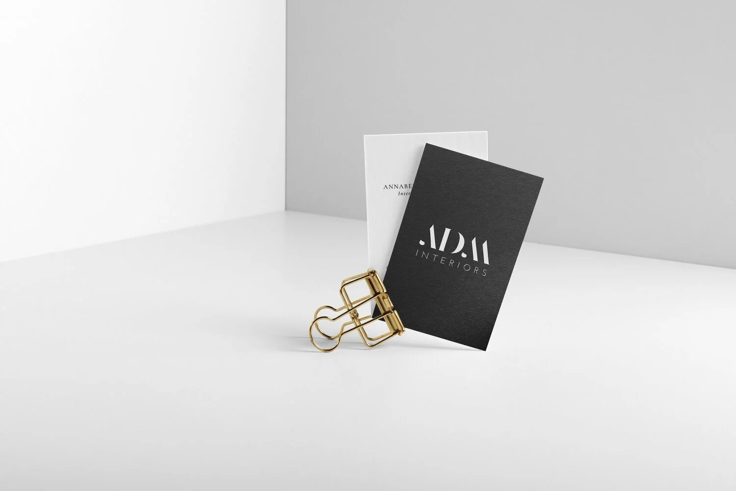 Two business cards, one black and one white, clipped together with a gold binder clip on a white surface against a gray background. The black card has the text 'ADM INTERIORS' in white.