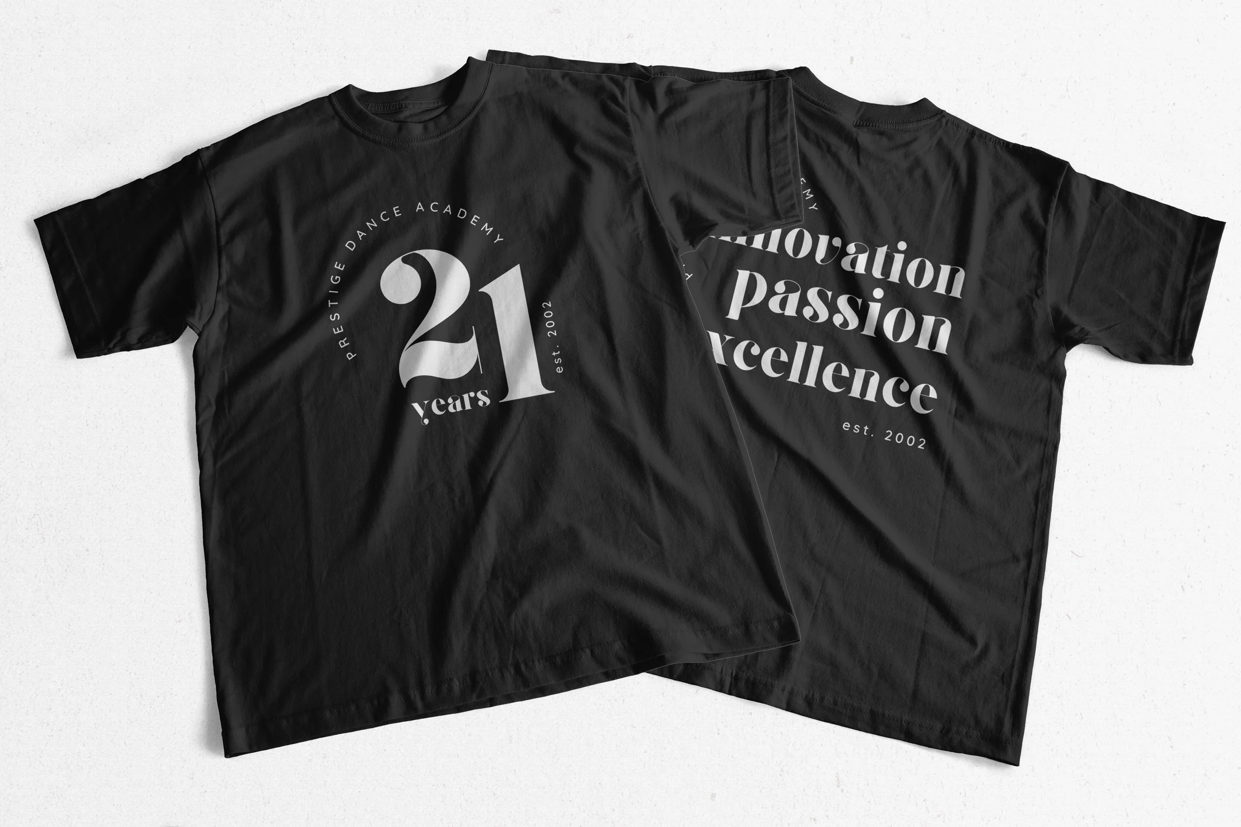 Two black T-shirts displaying "Prestige Dance Academy 21 years" with "Innovation Passion Excellence est. 2002" printed on them.