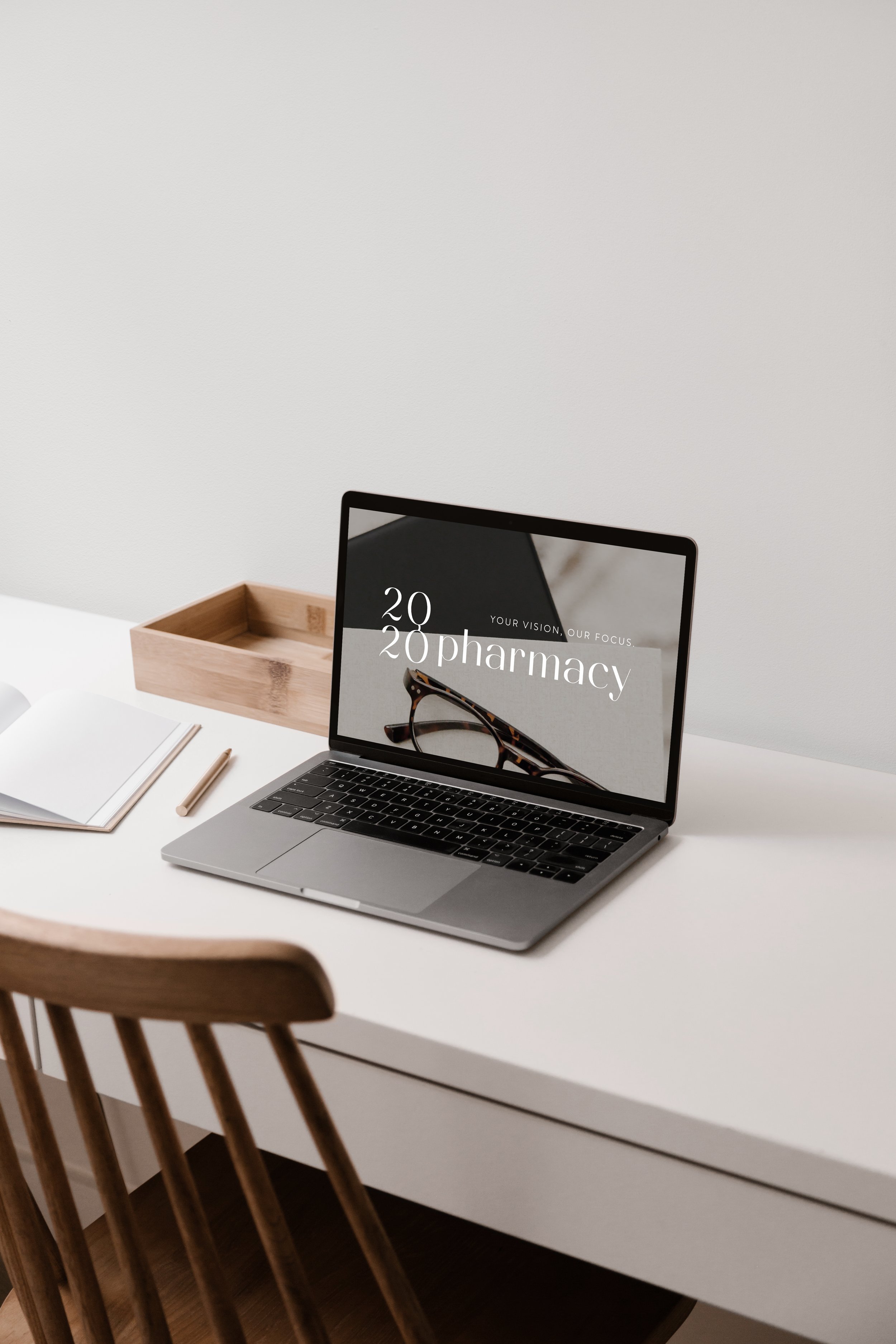 Minimalist workspace with a laptop displaying "20/20 pharmacy," a notebook, pencil, and wooden box on a white desk.
