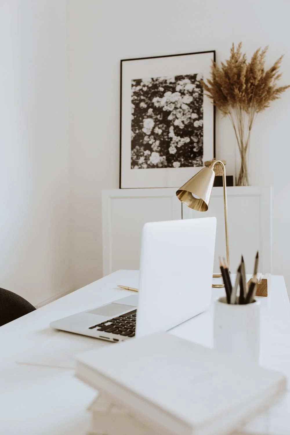 Minimalist home office with a laptop, gold desk lamp, potted pencils, books, black and white floral artwork, and dried plants on a white desk.