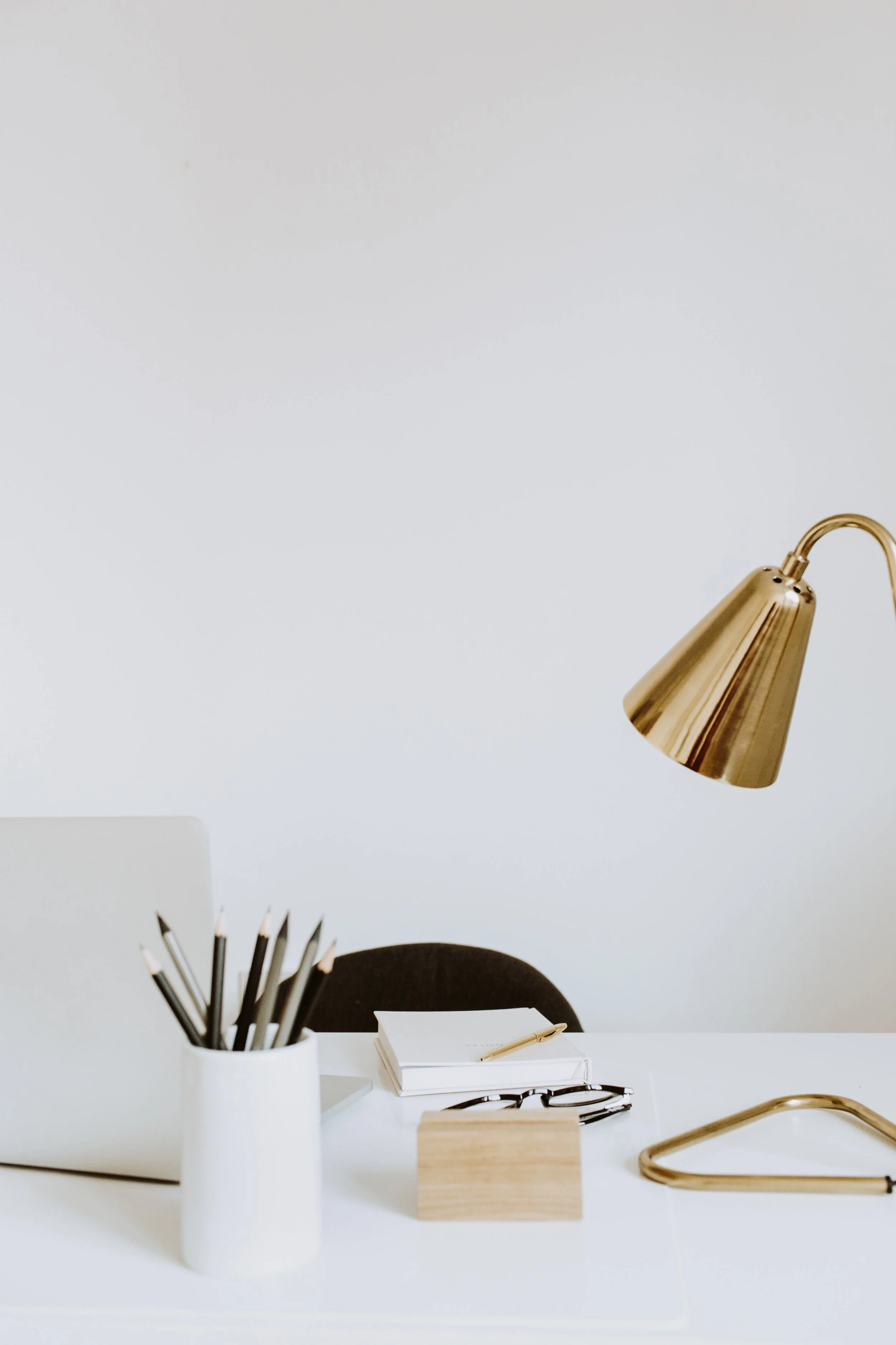Minimalist desk setup with gold lamp, pencils in white holder, stacked notebooks, a pen, glasses, and a laptop.