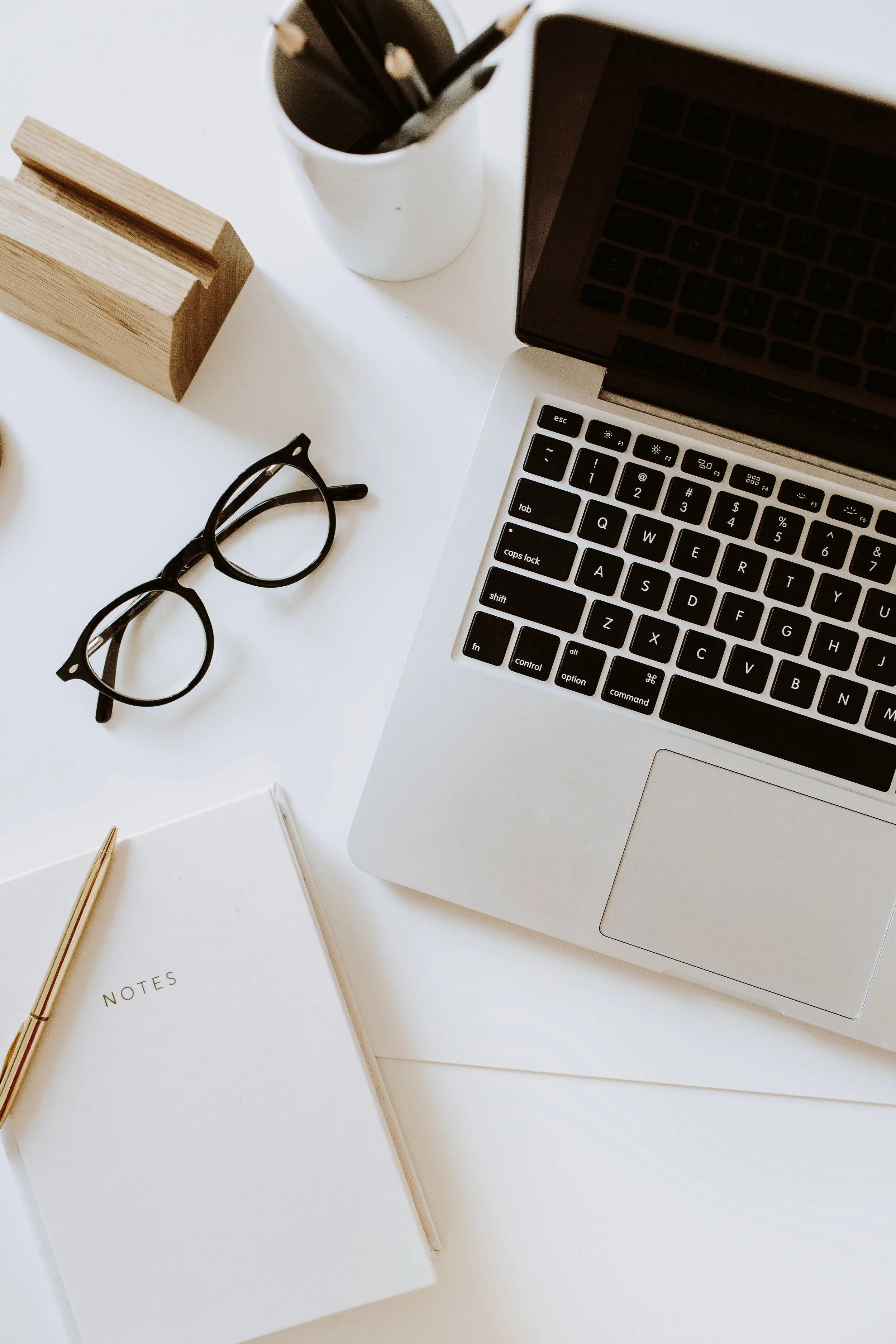 Laptop with black keyboard, glasses, white notebook labeled 'Notes,' and gold pen on white desk with wooden stand and cup of pencils.
