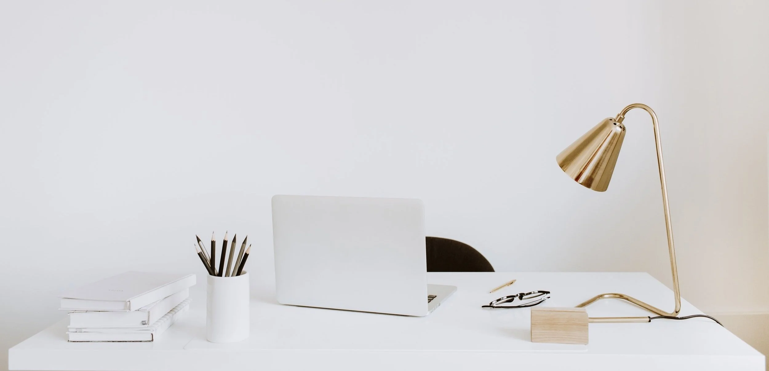 Minimalist white desk setup with a closed laptop, gold desk lamp, stack of books, pencil holder with pencils, and a pair of eyeglasses.