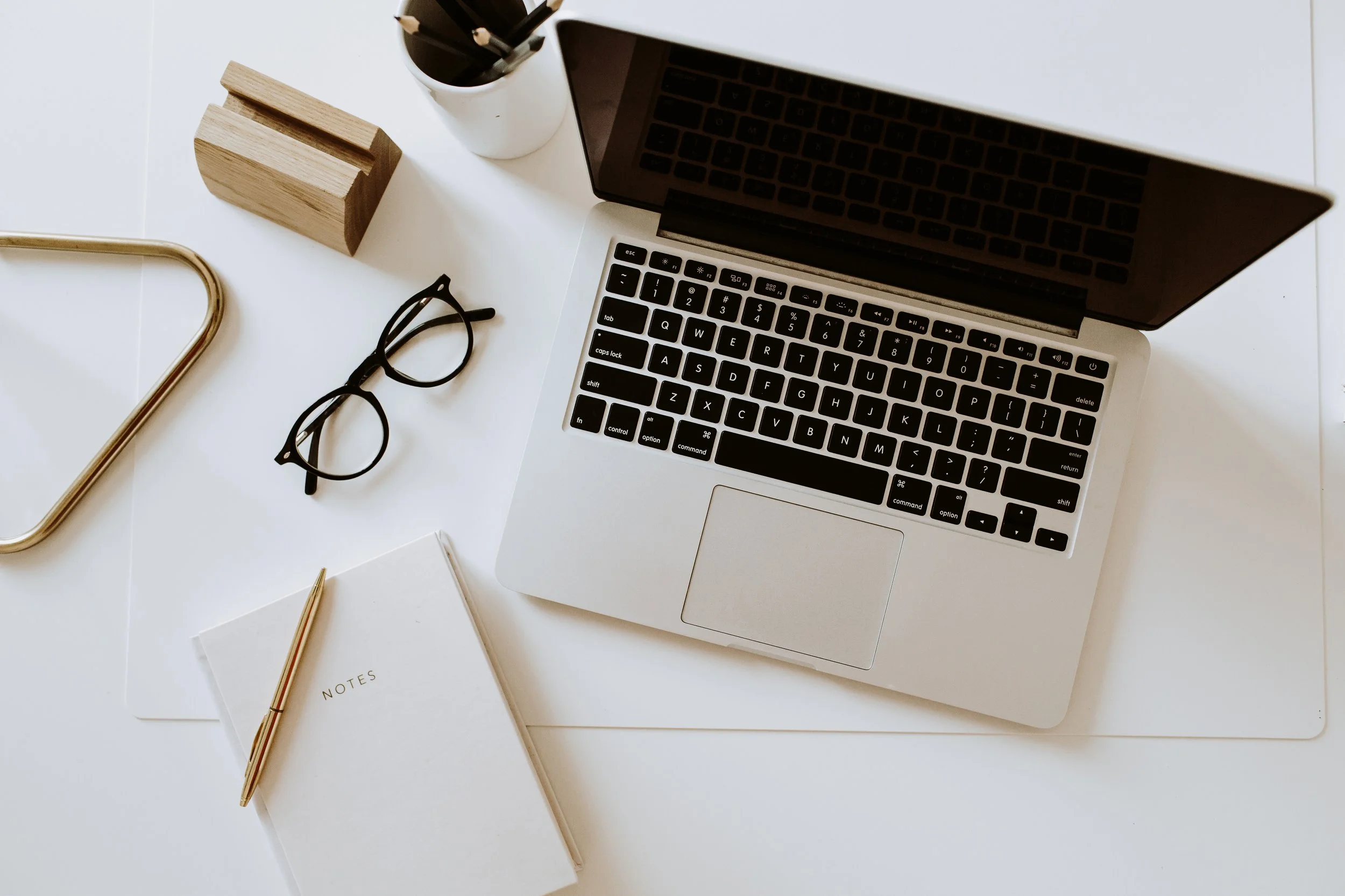Overhead view of a laptop, eyeglasses, notebook with pen, and a pencil holder on a white desk.