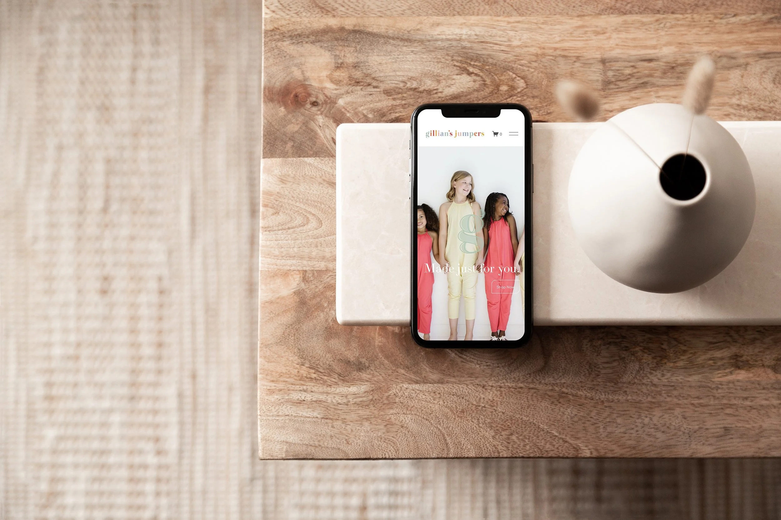 Smartphone on wooden table displaying a fashion website with images of children in colorful clothing, beside a white vase.