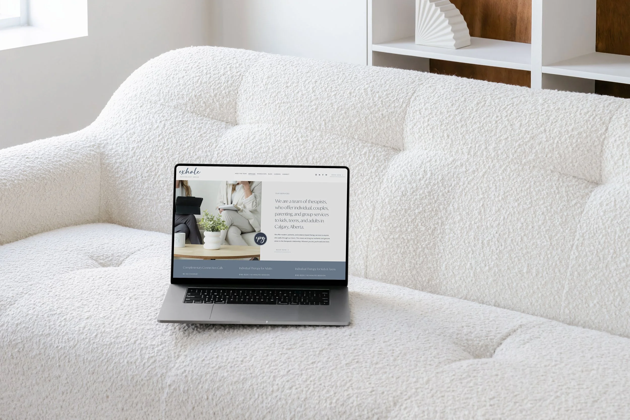 Laptop on a white textured couch displaying a therapy center website with text about services for kids, teens, and adults in Calgary, Alberta. White shelving with a decorative item is visible in the background.