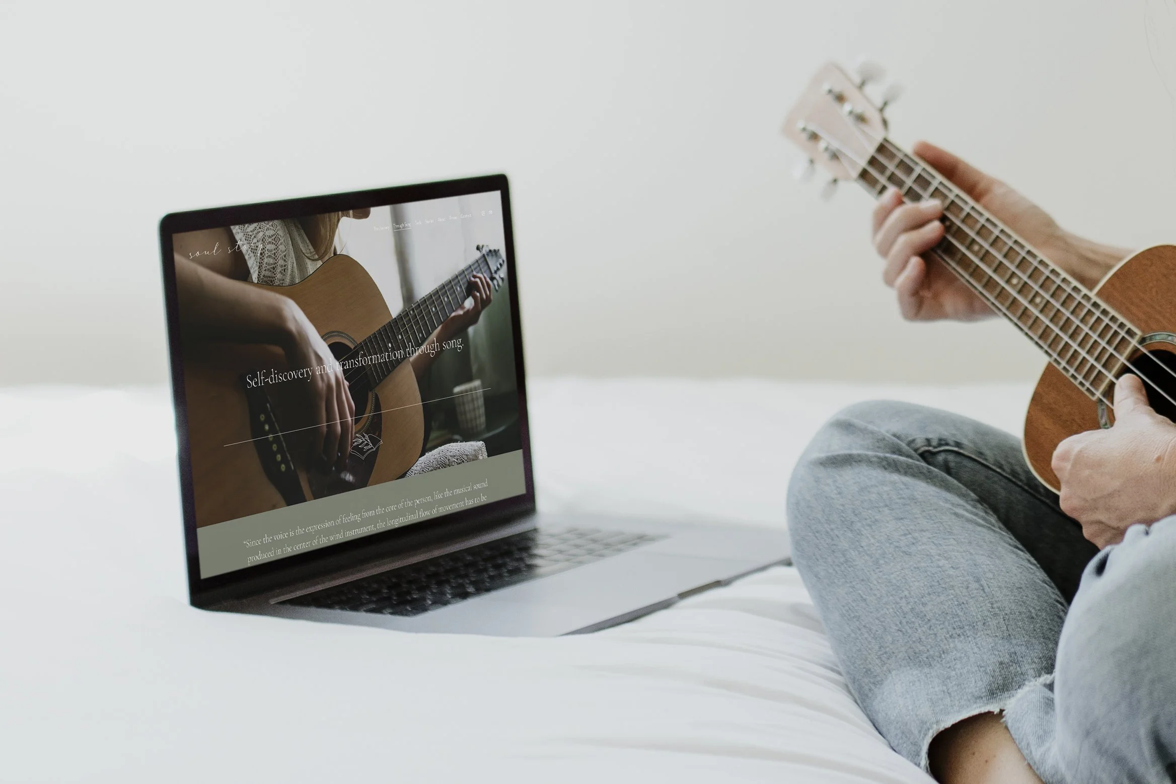 Person playing a ukulele while sitting on a bed next to an open laptop showing a website about self-discovery through music.