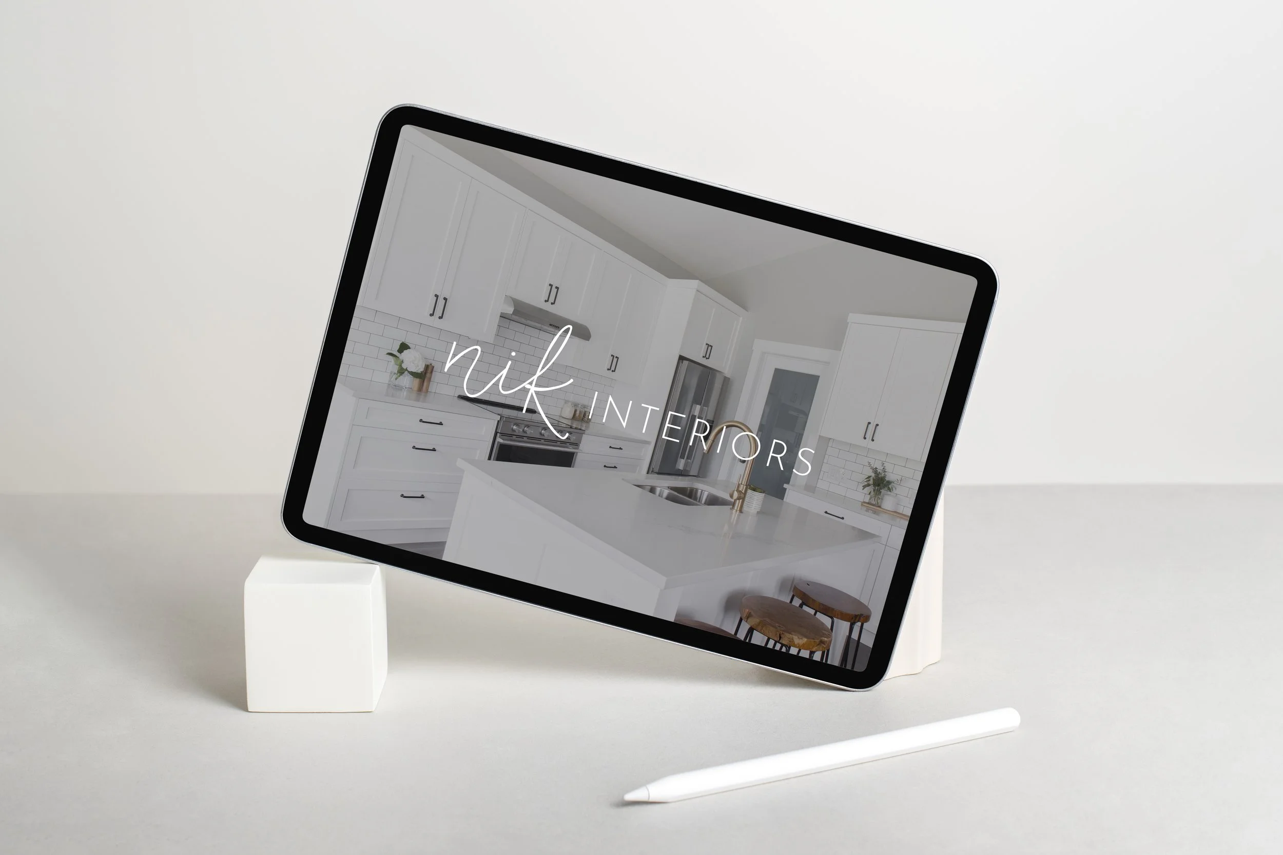 Tablet displaying a kitchen interior design with white cabinets and modern decor, labeled 'nik INTERIORS', on a minimalist surface with a stylus.