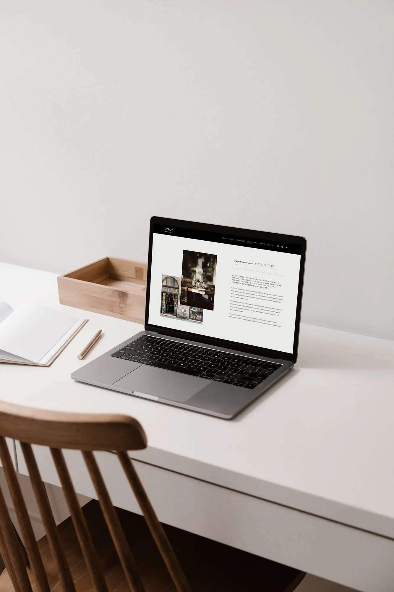 Open laptop on a white desk next to a wooden organizer, pencil, and notebook, with a wooden chair in front.