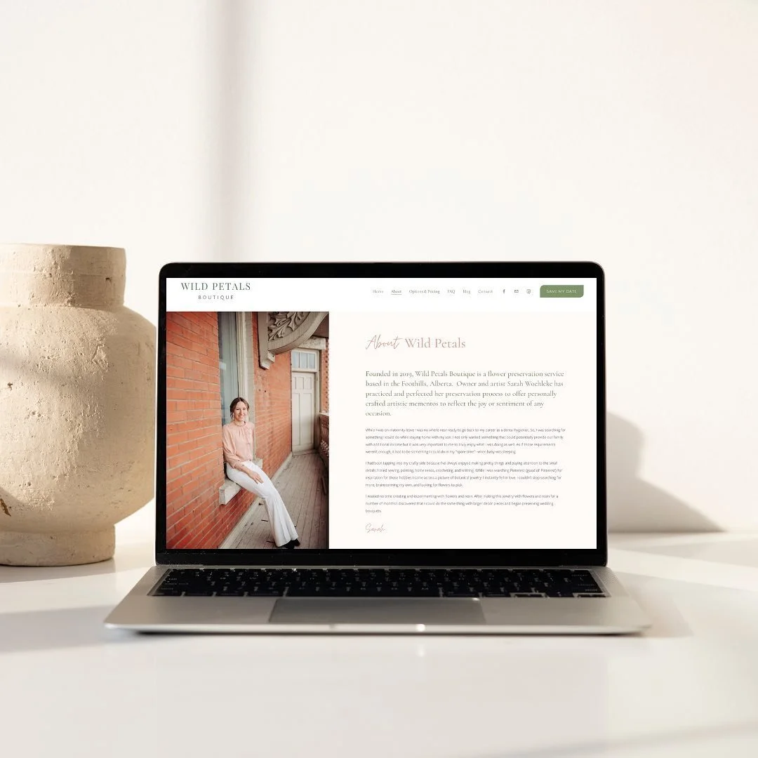 Laptop displaying a website with an 'About Wild Petals' page featuring a person sitting against a red brick wall. A large vase is next to the laptop on the white surface.