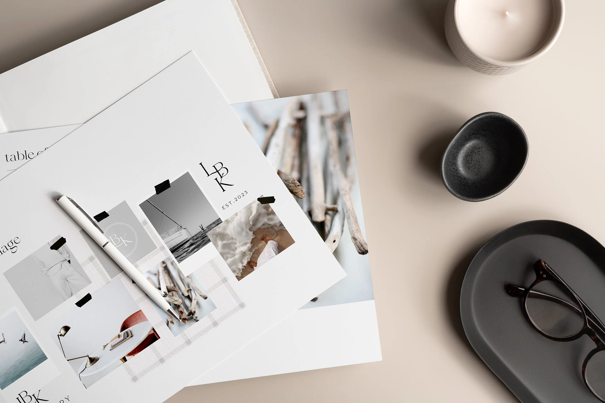 Flat lay of design mood board with photos and branding elements, a candle, small black dish, gray tray with eyeglasses on a light beige surface.