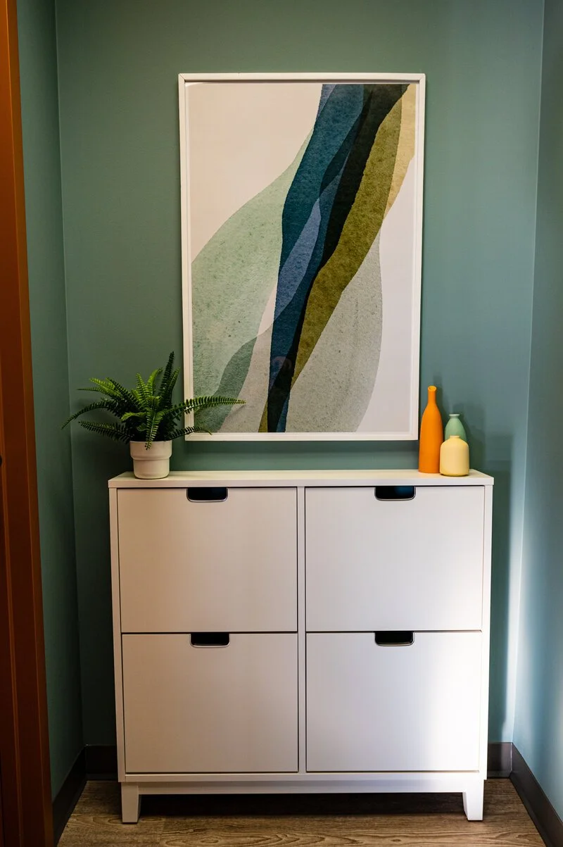 A white storage cabinet with four drawers is placed against a teal wall. On top of the cabinet, there is a potted fern, and three decorative vases in yellow, green, and orange. Behind the cabinet, a large framed abstract art print with curved shapes 