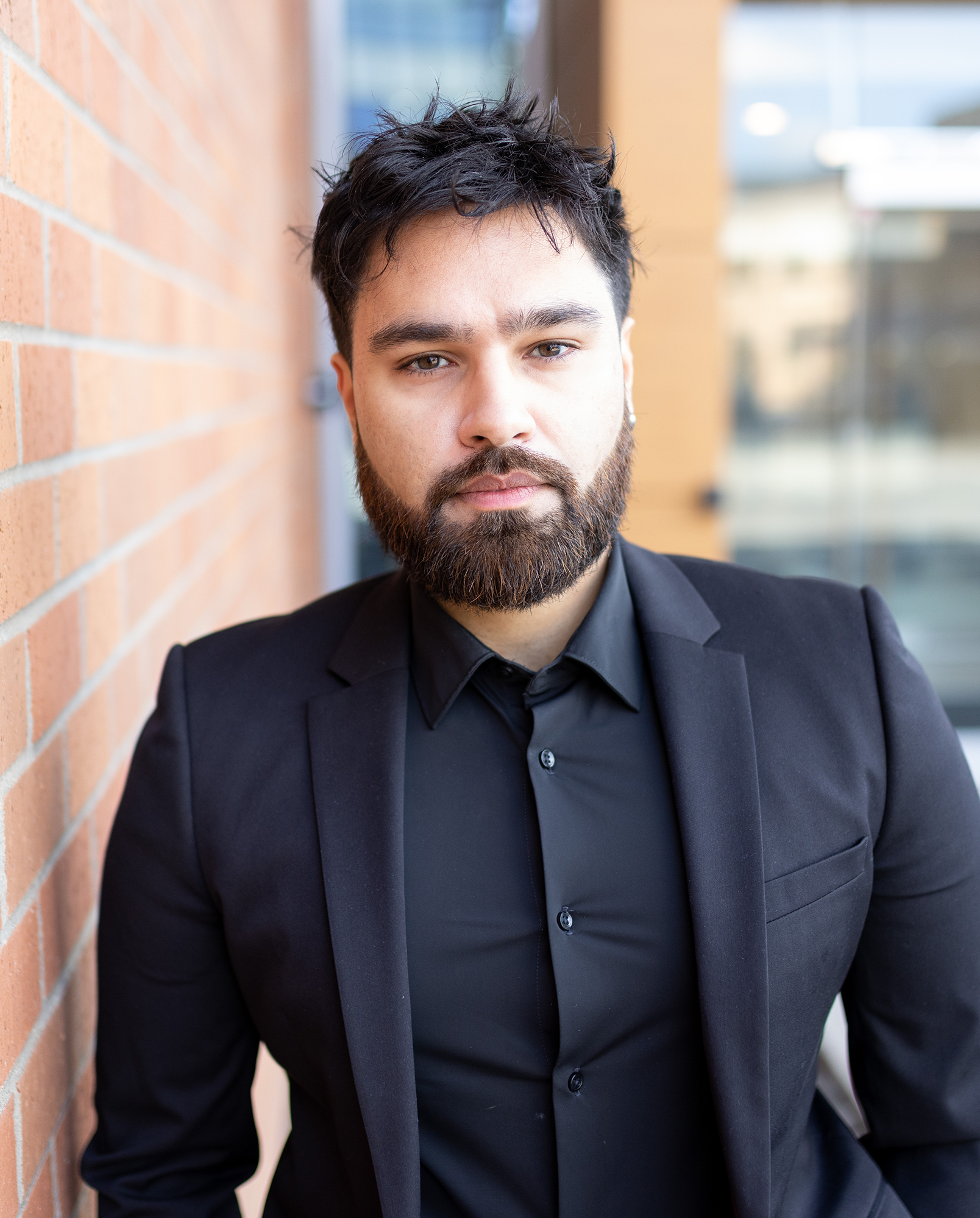 A man with dark hair and a beard wearing a black suit, standing outdoors against a background of a brick wall and glass windows.